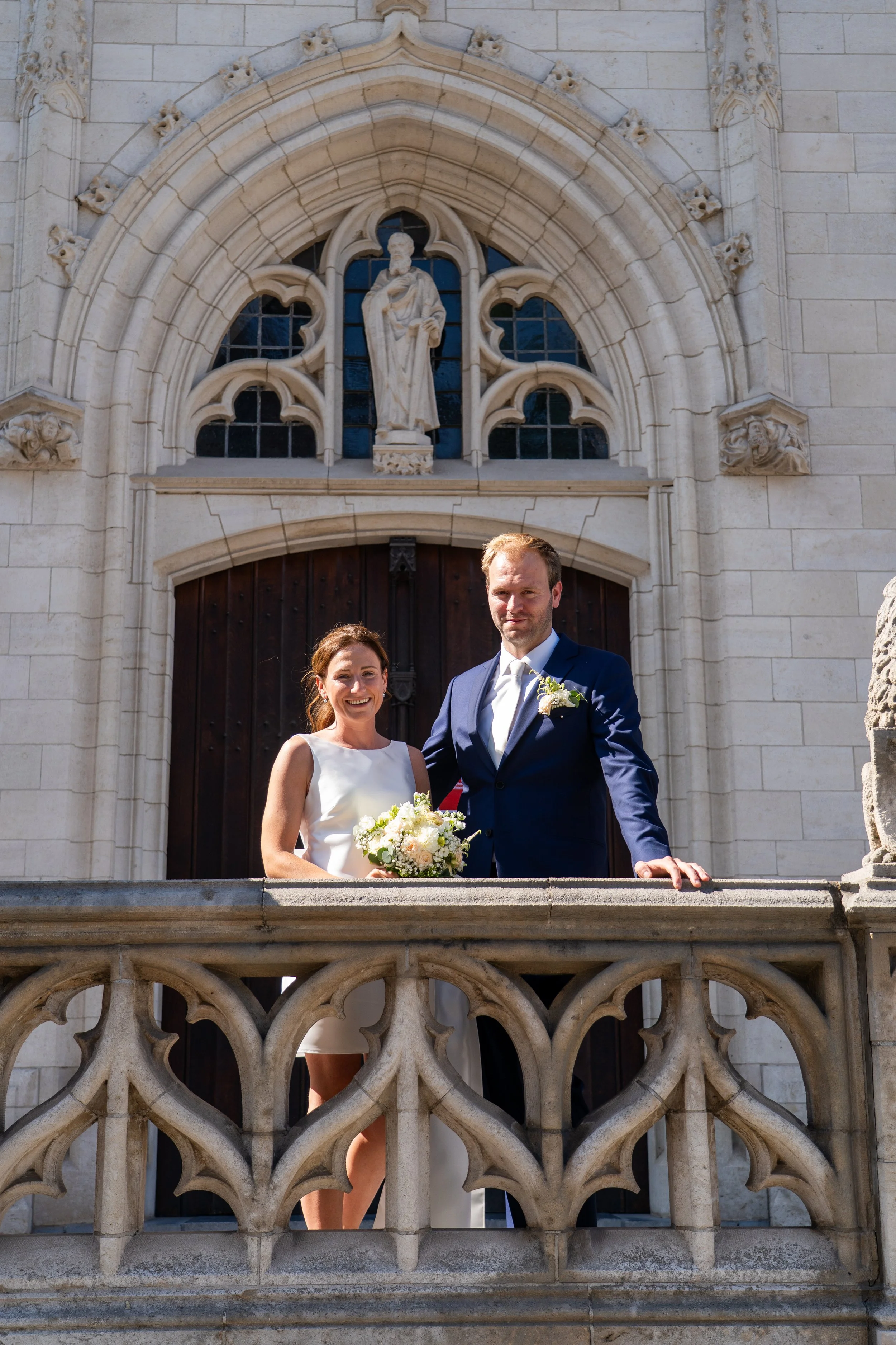 Een getrouwde stel op een balkon voor een kerk, zij houdt een bloemenboeket vast terwijl zij glimlacht, hij draagt een blauwe tuxedo met een witte blouse en bloemcorsage. Op de achtergrond een grote openslaande deur en een stenen gevel met beeldhouwwerken en een standbeeld boven de deur.