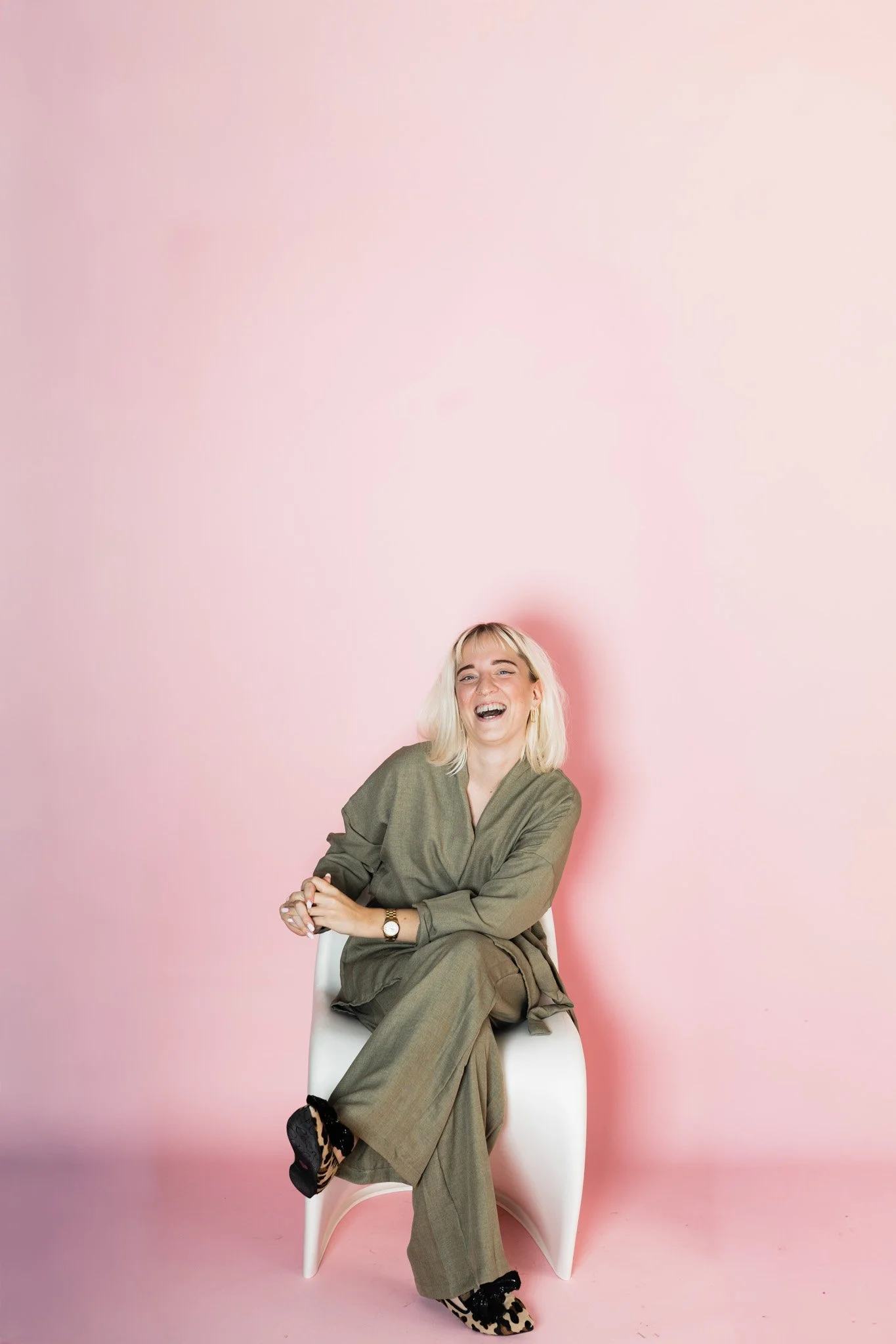 Een lachende vrouw met blond haar, zittend op een witte stoel, gekleed in een groene outfit, voor een roze achtergrond.