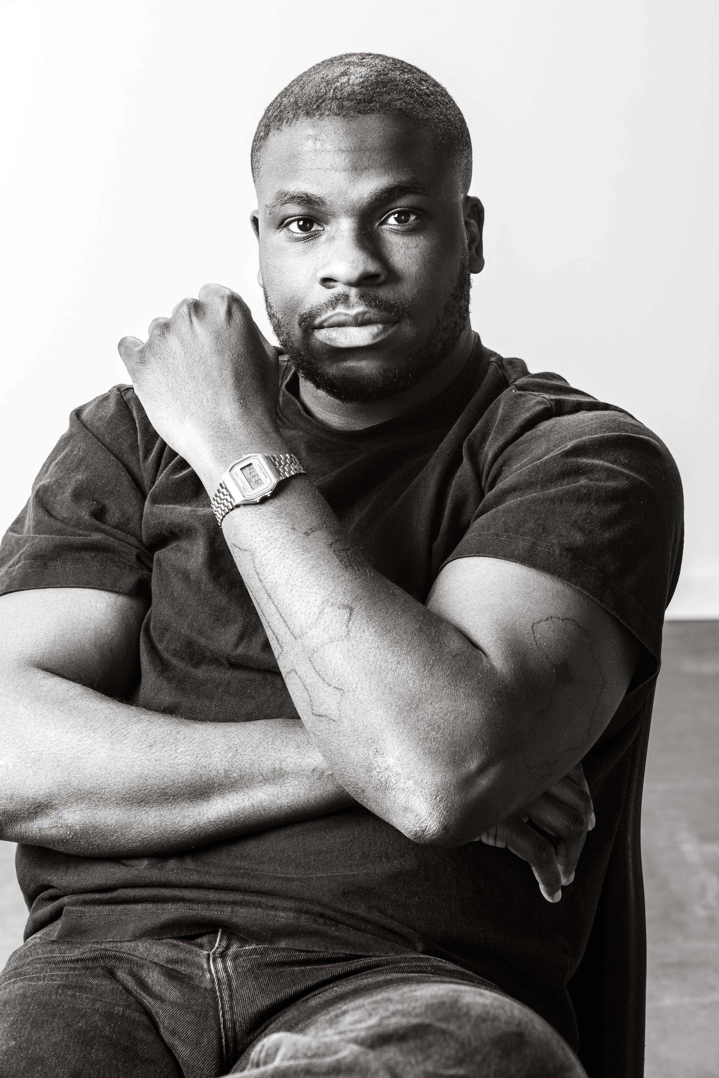 Zwart-witte foto van een man met een kort kapsel, een baard en tattoos op zijn onderarm, die een comfortabele pose aanneemt, met één arm over zijn borst en een draait draait de polshorloge.