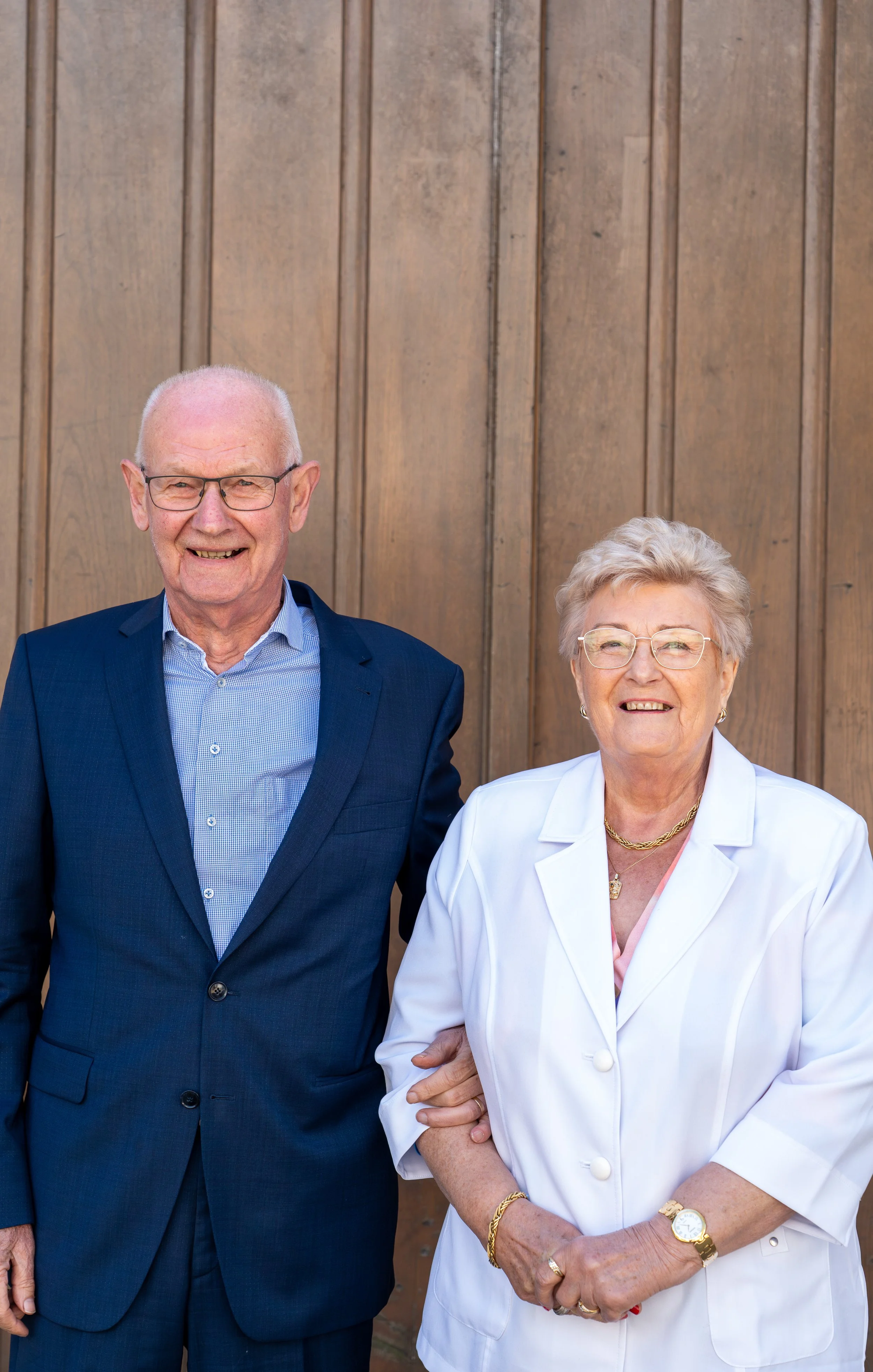 Een oudere man en vrouw staan samen voor een houten achtergrond, zij glimlachen en dragen formele kleding, de man draagt een blauw pak en de vrouw een witte jas.