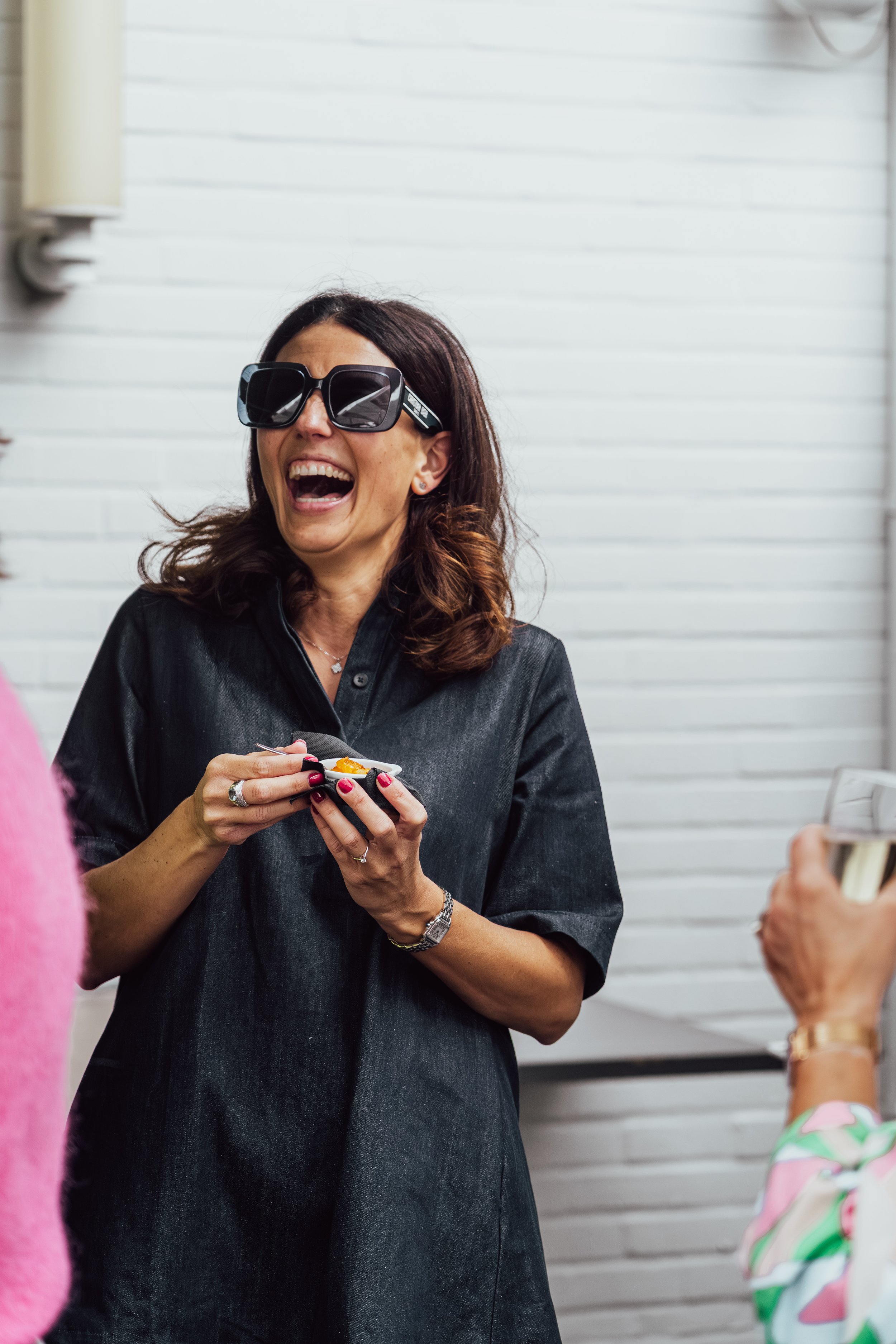 Vrolijke vrouw met grote zwart zonnebril en zwarte jurk, lachend en eten vasthoudend in een binnenruimte met witte muren.