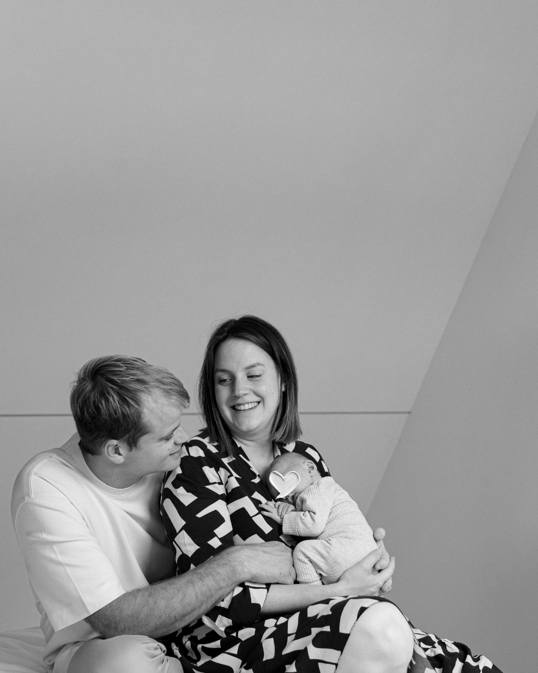 Gezicht van een vrouw die haar pasgeboren baby vasthoudt, terwijl haar partner erbij zit en naar hen kijkt, in zwart-wit.