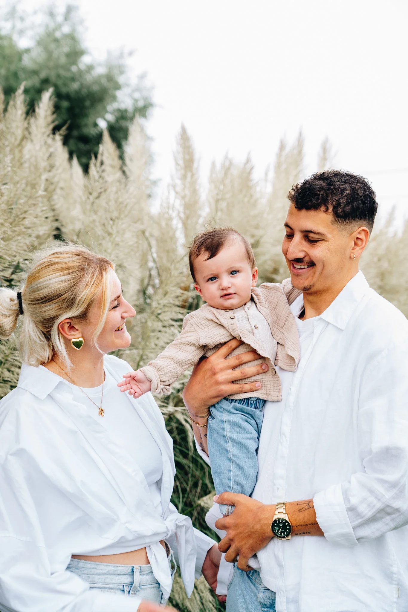 Een gezin met een man, een vrouw en een kind staat buiten in een veld met hoge, pluizige planten. De man houdt het kind op en kijkt naar haar met een glimlach, terwijl de vrouw naar het kind lacht.