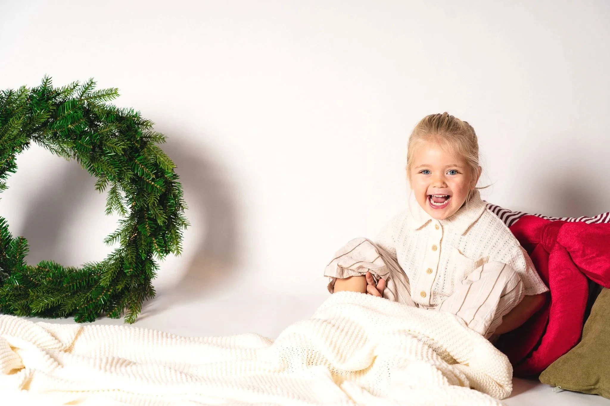 Een lachend meisje dat op een bed ligt met kussens, naast een groene kerstkrans, in een lichte kamer.