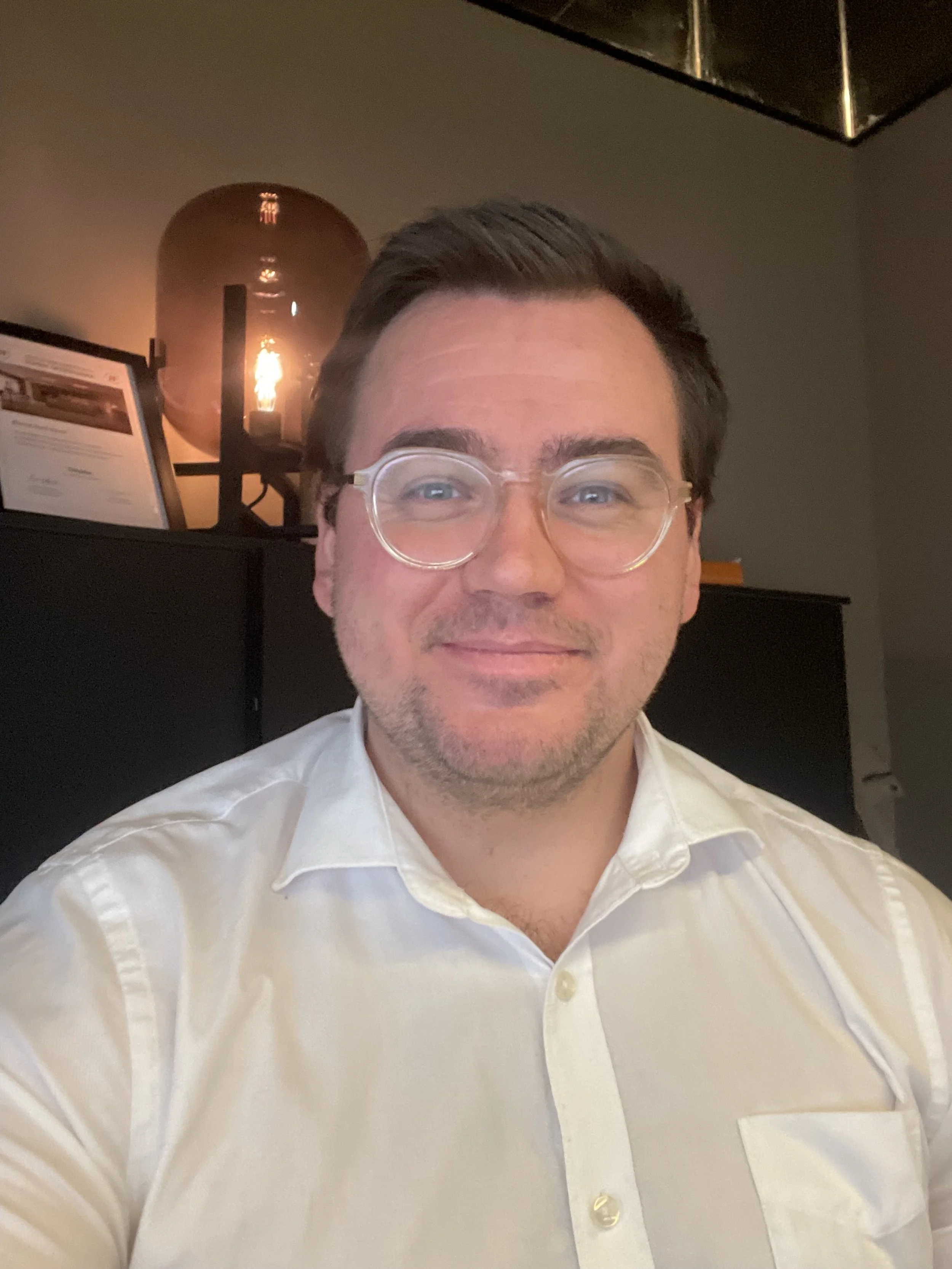 A man with glasses wearing a white dress shirt, sitting in a room with a lamp and framed certificate in the background.