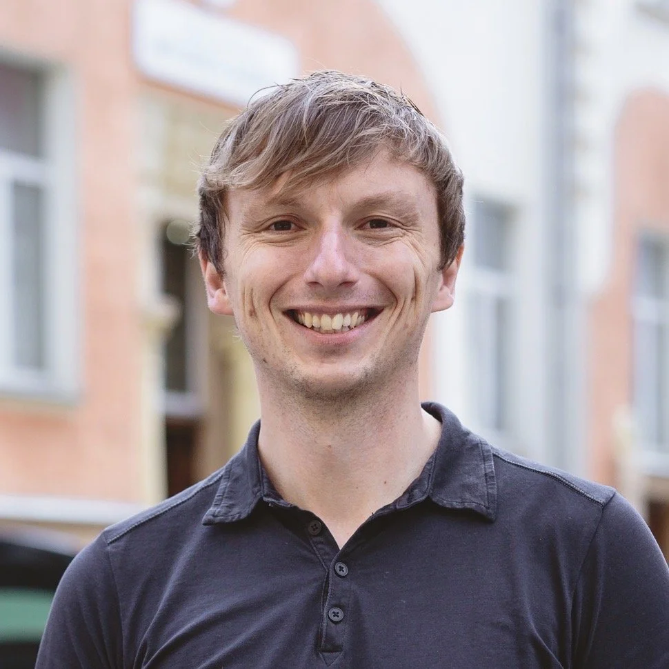 A young man with light brown hair, smiling, wearing a black collared shirt, standing outdoors in a residential area with brick buildings in the background.