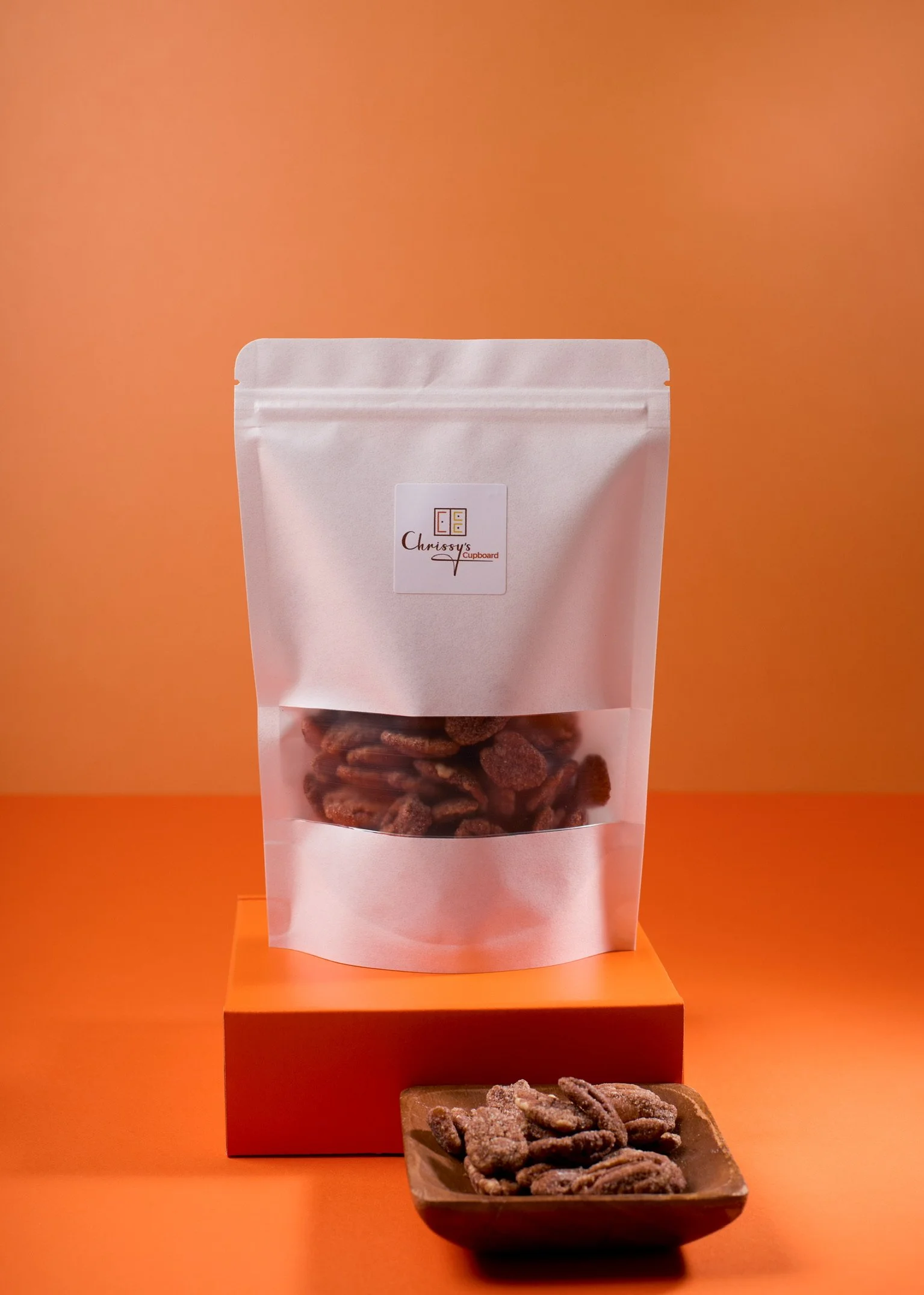 Small tray of pecans sitting in front of white bag of roasted pecans on top of orange box