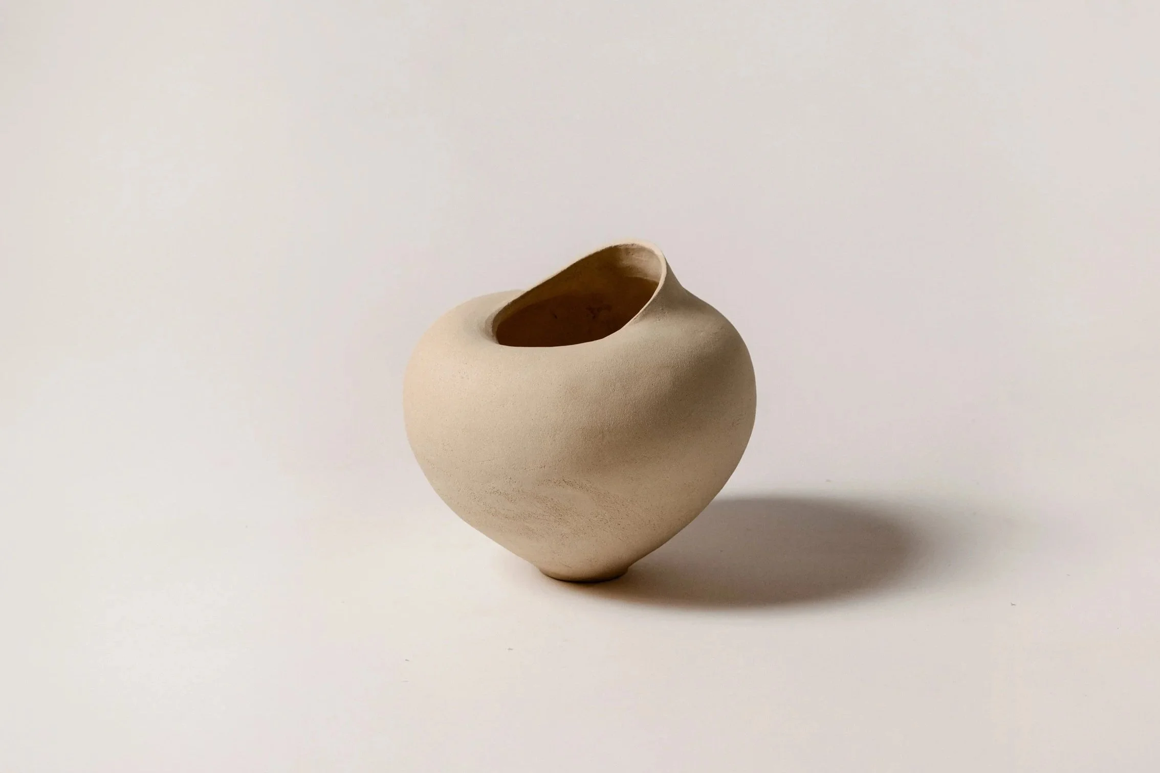 A beige ceramic vase with a round shape and an opening at the top, placed on a white surface with a plain white background.