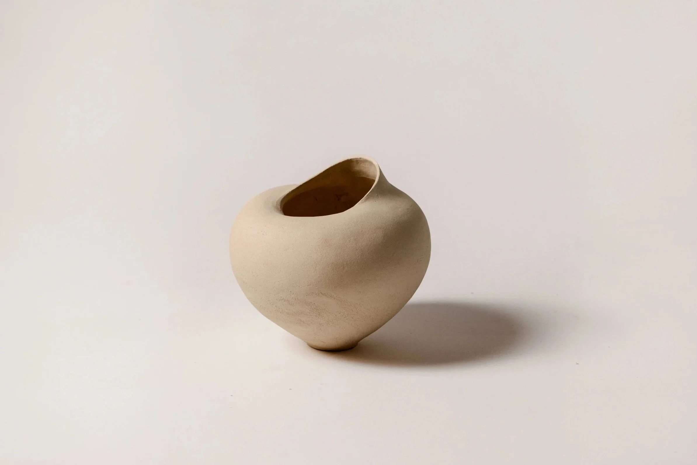 A smooth, beige ceramic vase with a rounded, asymmetrical shape and a large opening at the top, placed on a white surface with a shadow cast on the right.