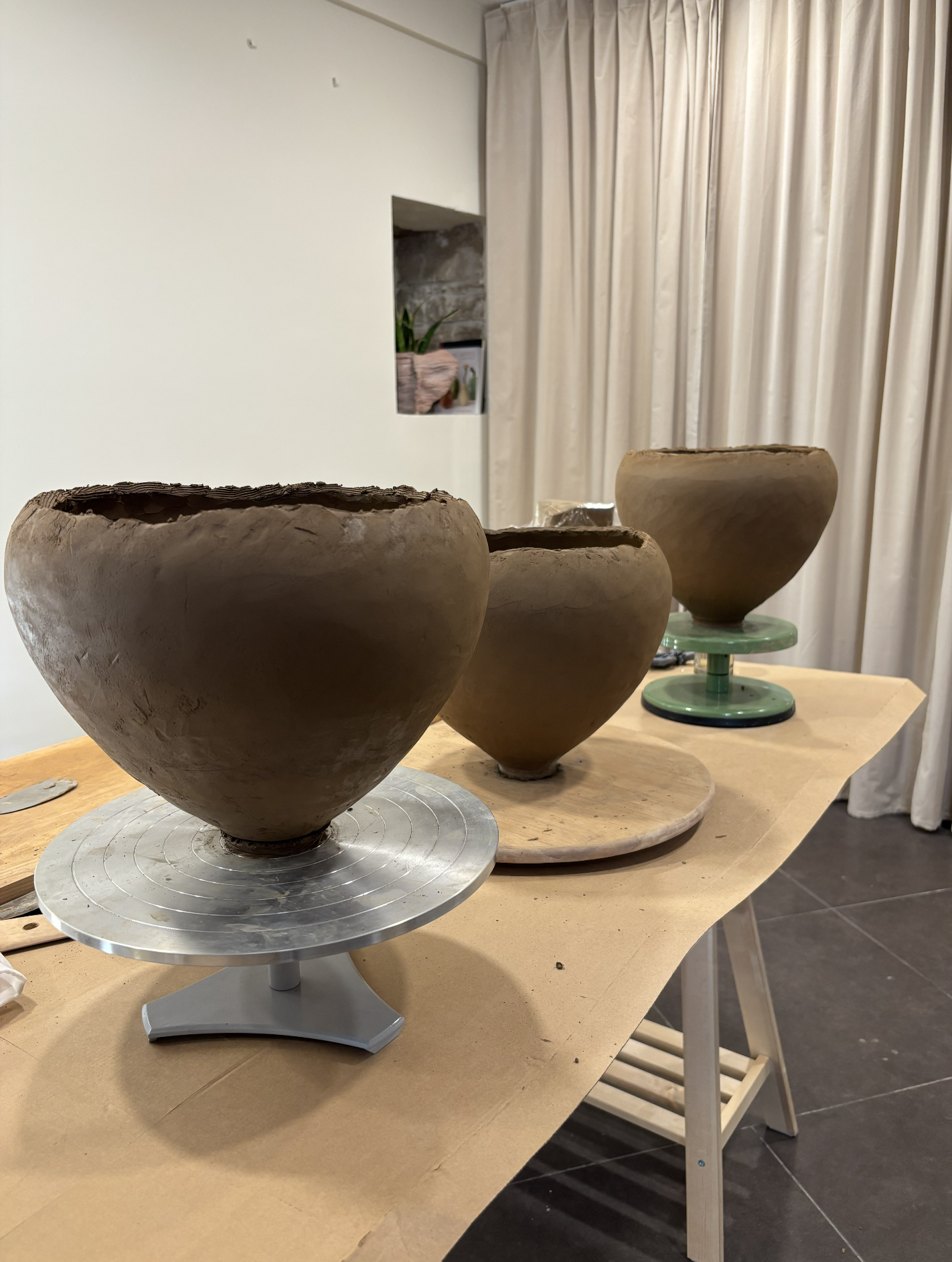 Studio image. Three large clay vessel placed on rotating stands on a wooden table, with a background of beige curtains and a white wall with a small opening.