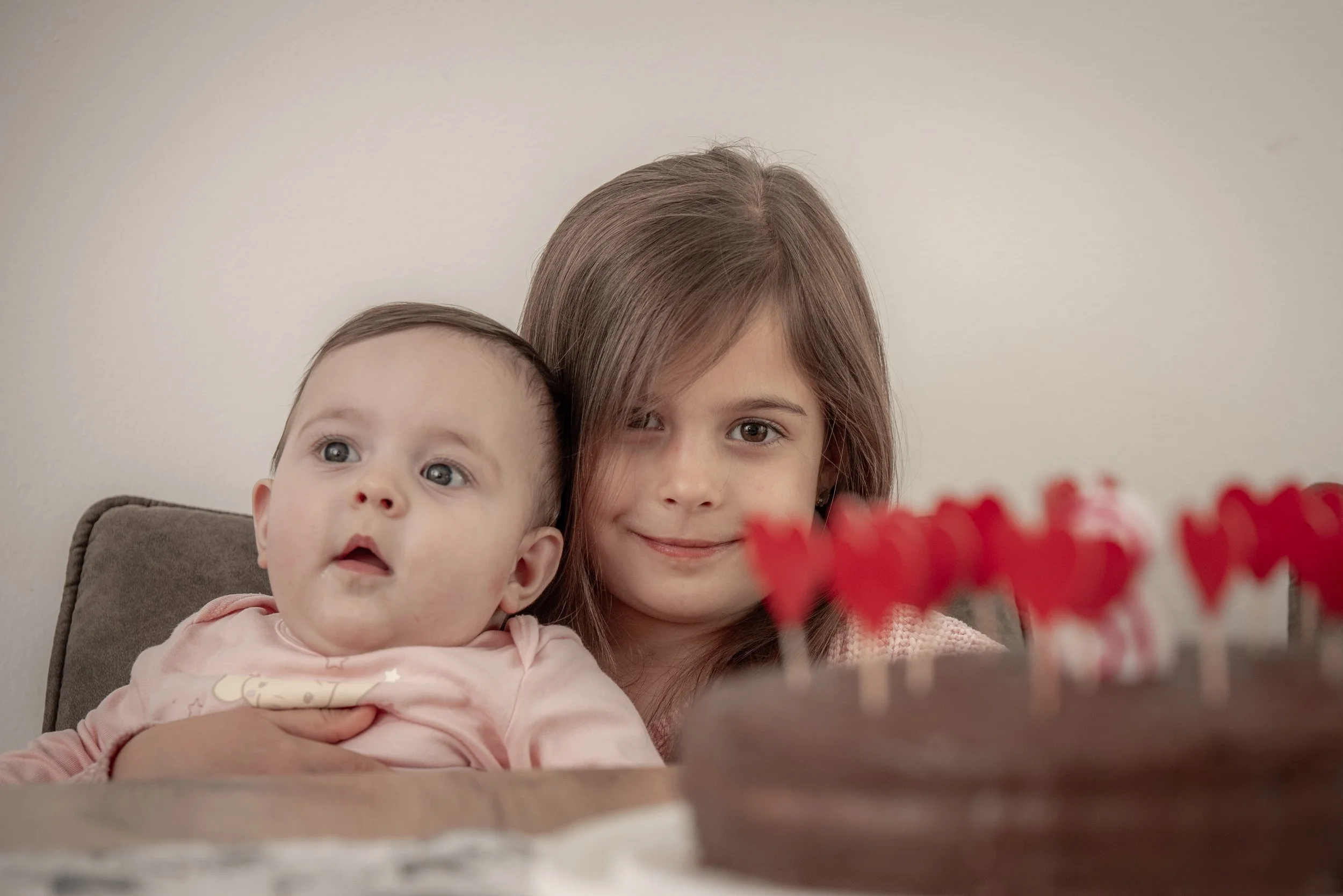 A young girl and a baby with a chocolate cake in the foreground, adorned with red heart decorations.