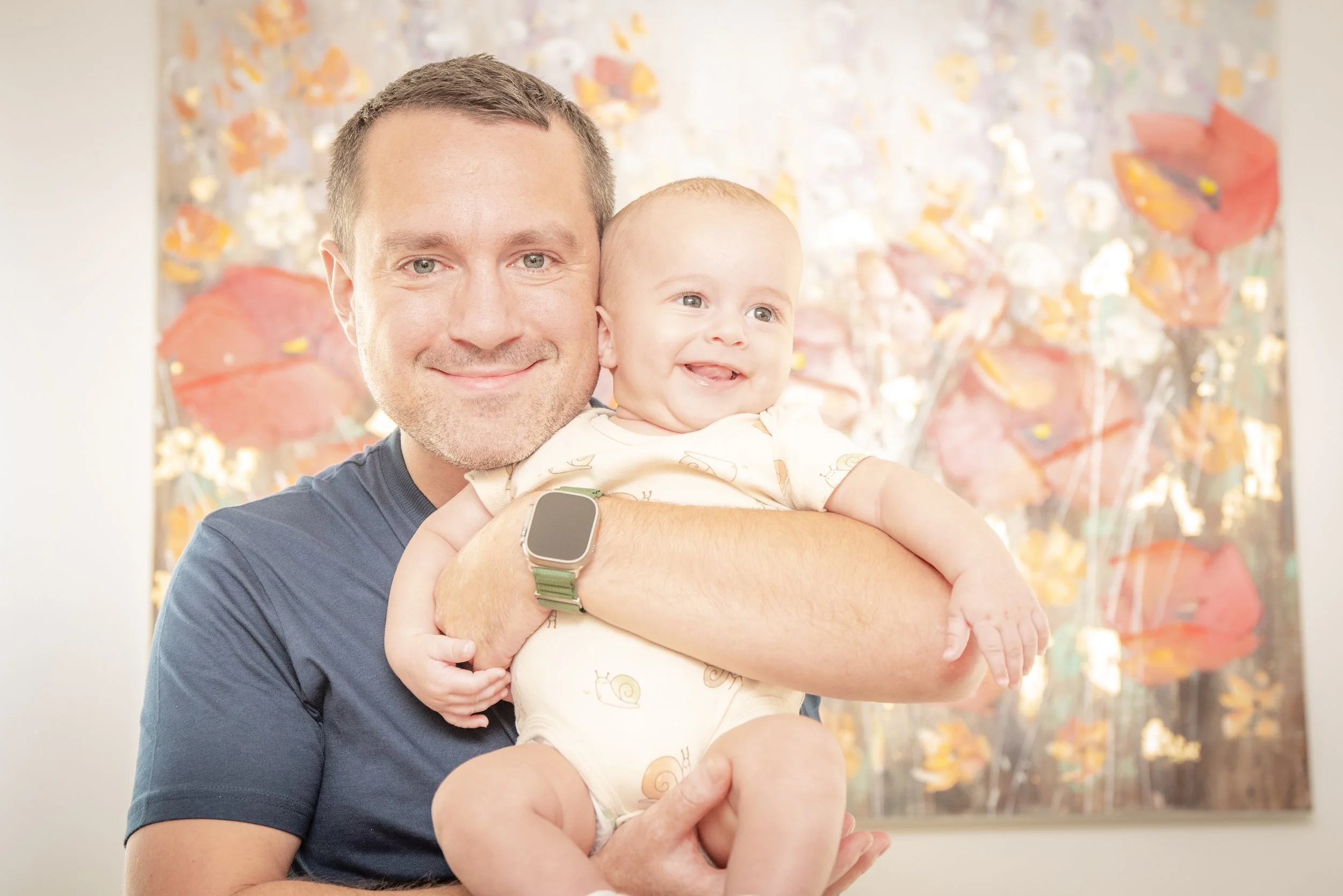 Man holding a smiling baby in front of a colorful floral painting.