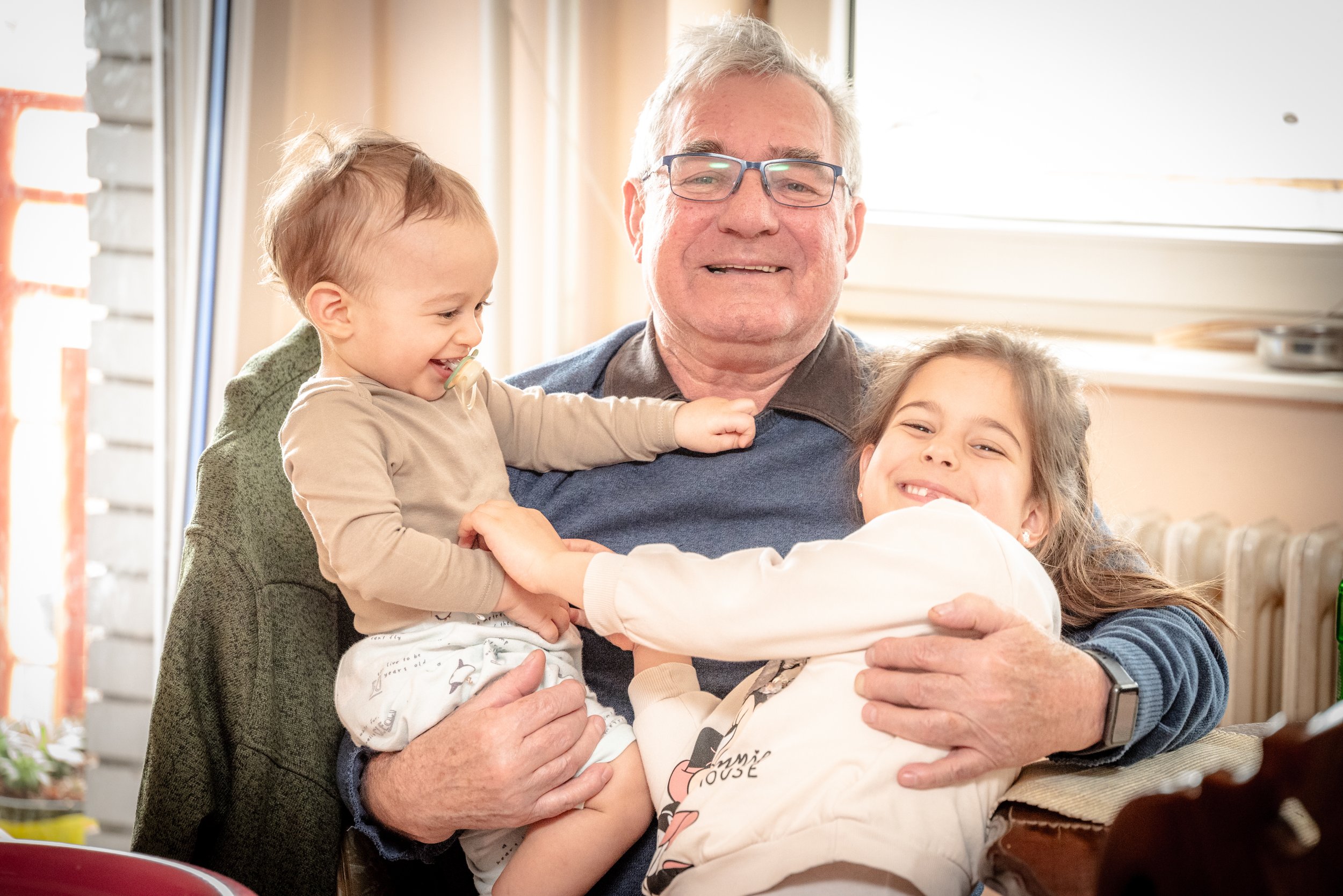 Grandfather smiling and holding two young children indoors, all appearing happy and playful.