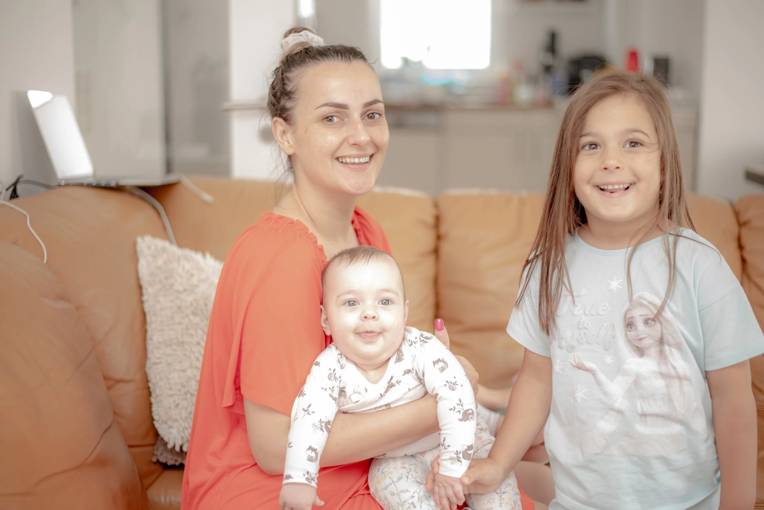 A woman holding a baby and sitting on a brown sofa next to a young girl wearing a light blue shirt, all smiling.