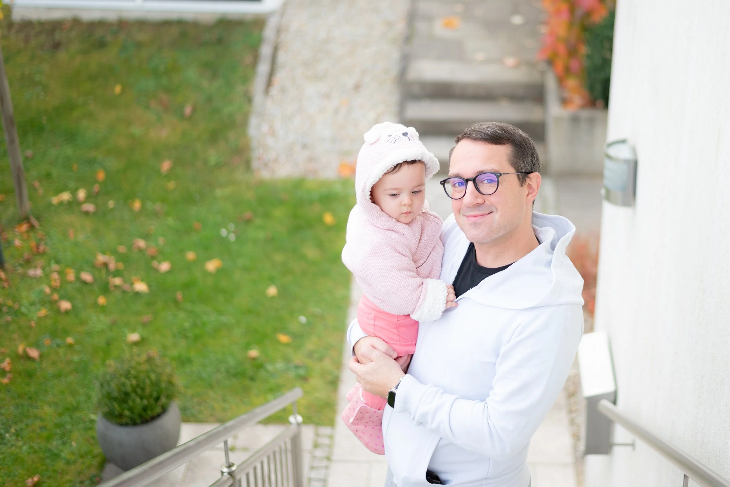 A man holding a baby on outdoor stairs, with grass and leaves in the background.