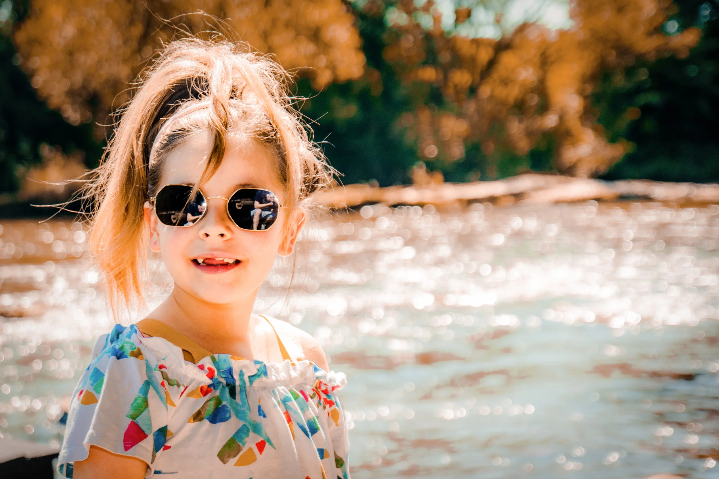 Young girl wearing sunglasses with trees and water in the background.