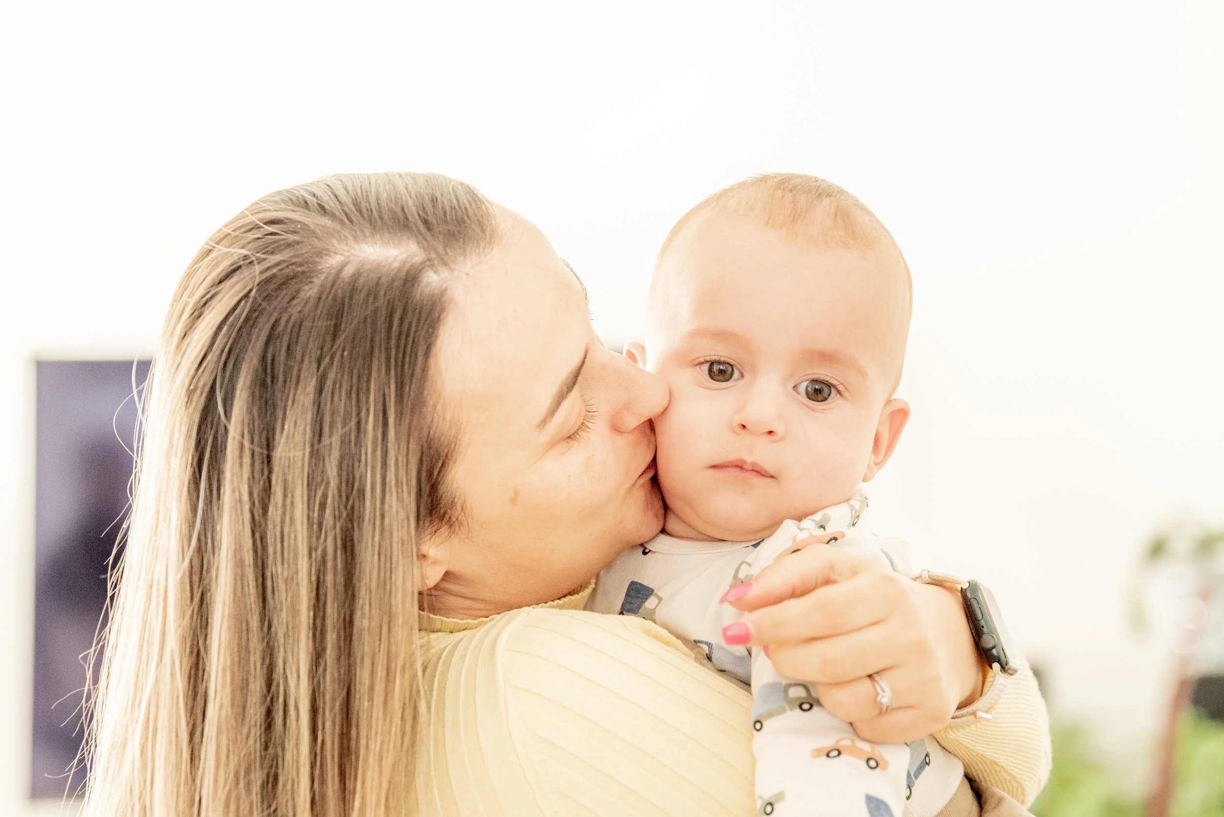 A woman holding and kissing a baby, both with fair skin. The baby is wearing a patterned outfit, and the woman has long hair and is wearing a light-colored top. She is also wearing a watch and a ring.
