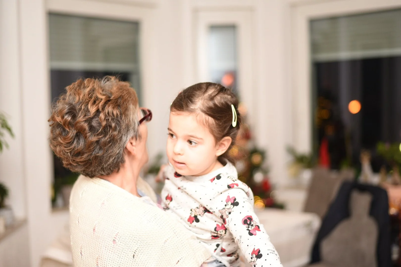 Elderly person holding a young child in a Minnie Mouse shirt, both indoors in a warmly lit room with furniture and plants visible.