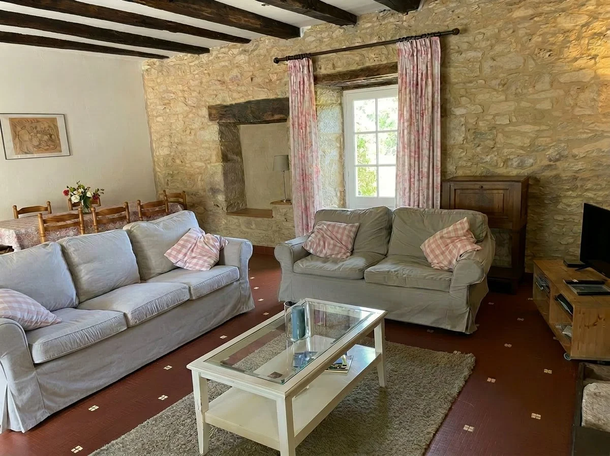 Living room with stone wall, wooden beams, two light-colored sofas with pink checkered pillows, a glass-top coffee table, a small TV on a wooden stand, a window with pink checkered curtains, and a dining area with a table and chairs in the background.