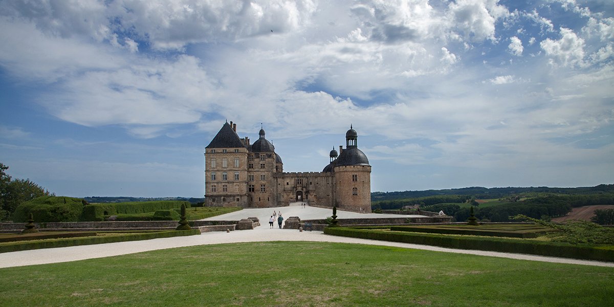 Château de Hautefort in the Dordogne with multiple towers and turrets, with a landscaped garden and pathway leading up to it, under a partly cloudy sky.