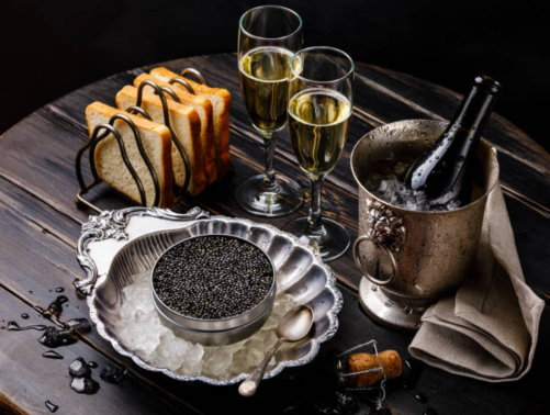 Charcuterie board with toasted bread slices drizzled with chocolate, two glasses of champagne, a champagne bucket with a bottle inside, a silver dish of caviar, and a small spoon, all arranged on a dark wooden table.
