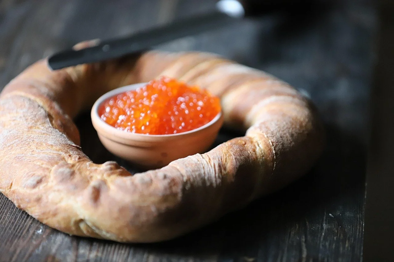 A bagel with a small bowl of orange salmon roe caviar on a dark wooden surface.