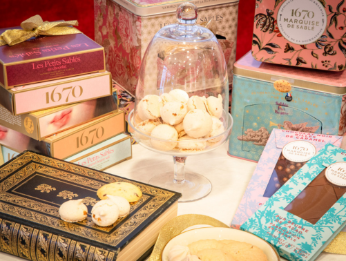 assortment of boxed chocolates, cookies, and macaroons on a display table with decorative packaging and glass dome cover.