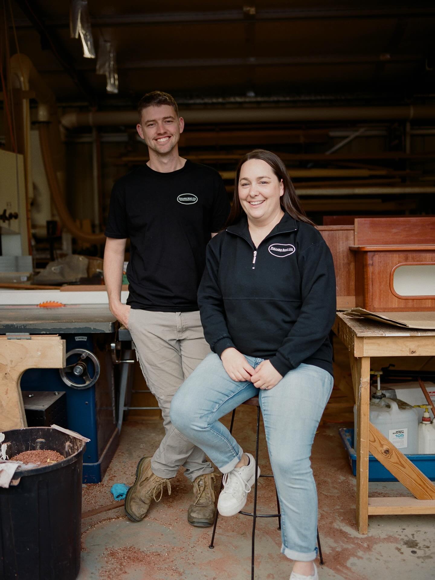 Two smiling people, a man and a woman, in a woodworking workshop, posing for the photo with woodworking tools and equipment in the background, both wearing black shirts with a logo.