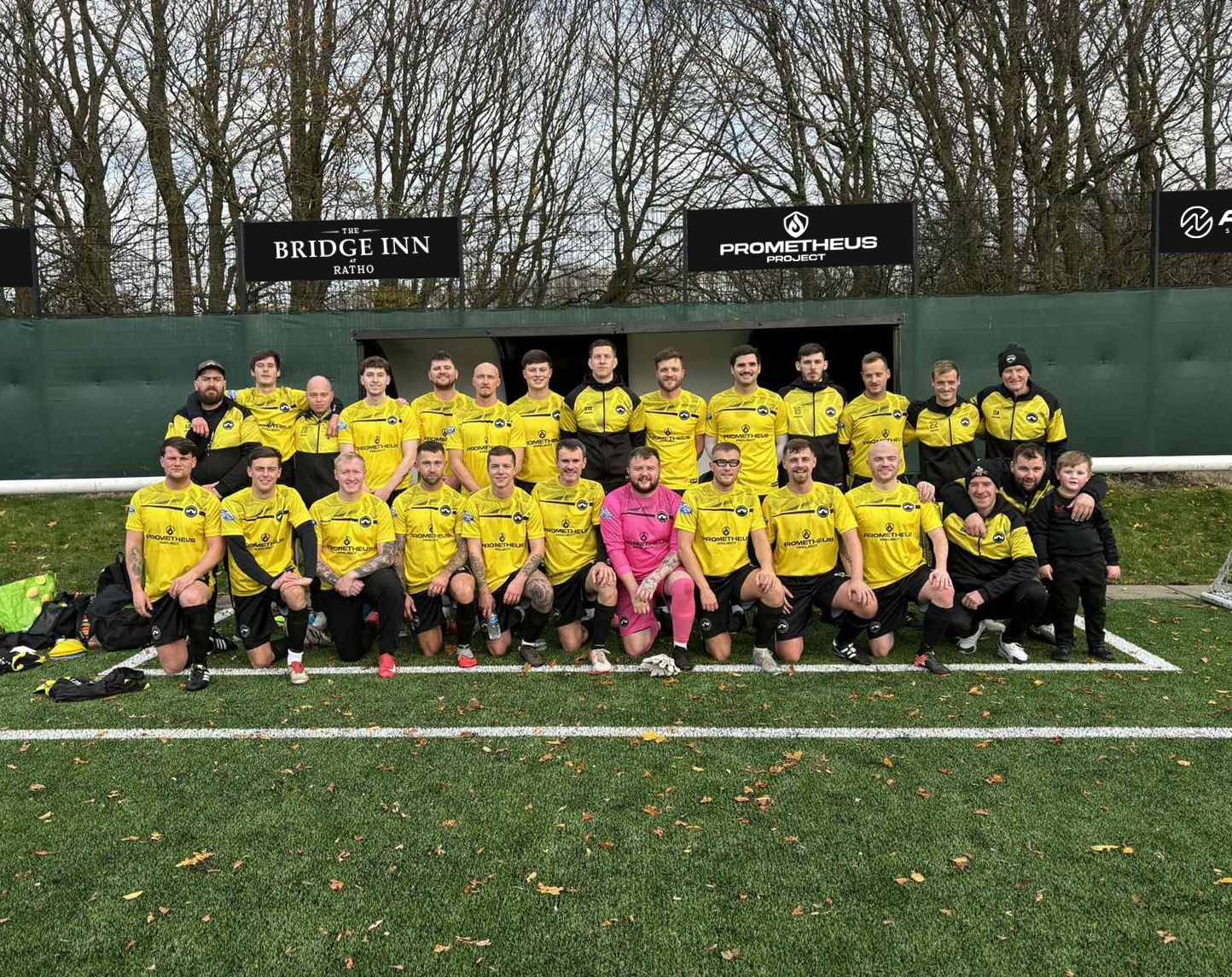 A soccer team wearing yellow and black uniforms posing on the field in front of banners, with some players kneeling in the front row and others standing behind them.