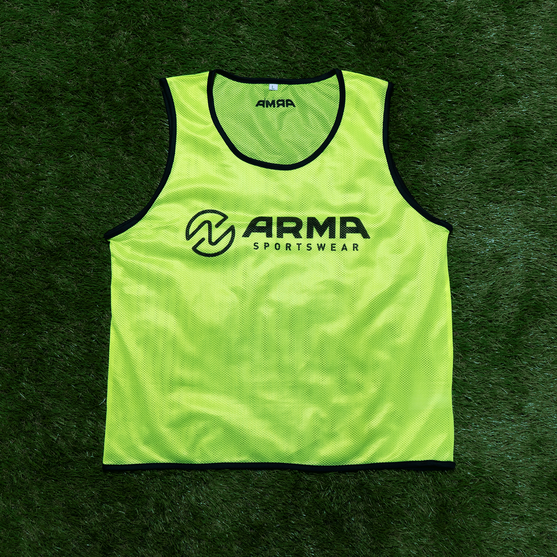 Bright yellow sports vest with black trim lying on green artificial grass. The vest features the logo and text "ARMA SPORTSWEAR" on the front.