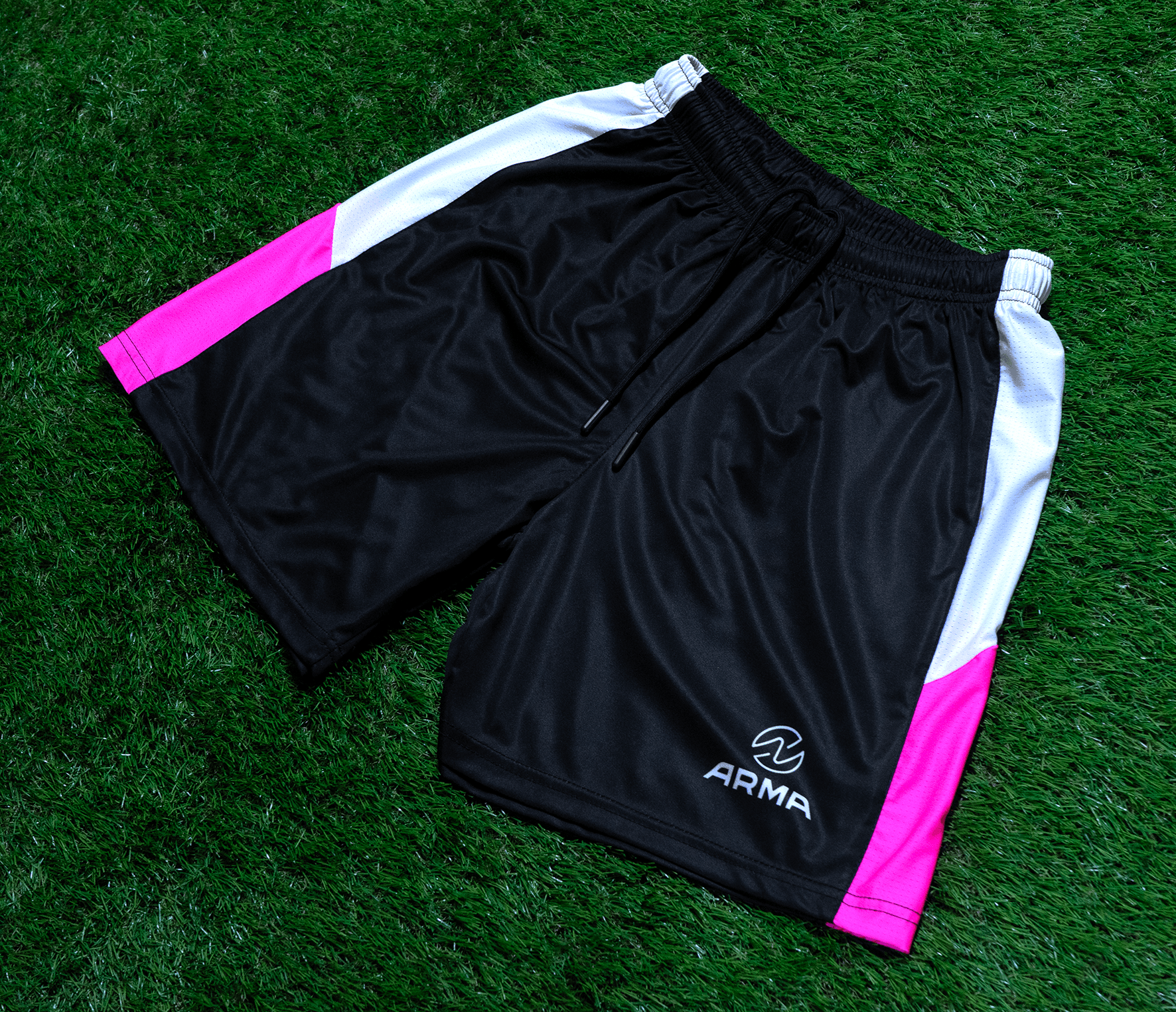 Black athletic shorts with white and pink side panels, placed on green grass, with a small logo reading 'ARMOUR' near the bottom.