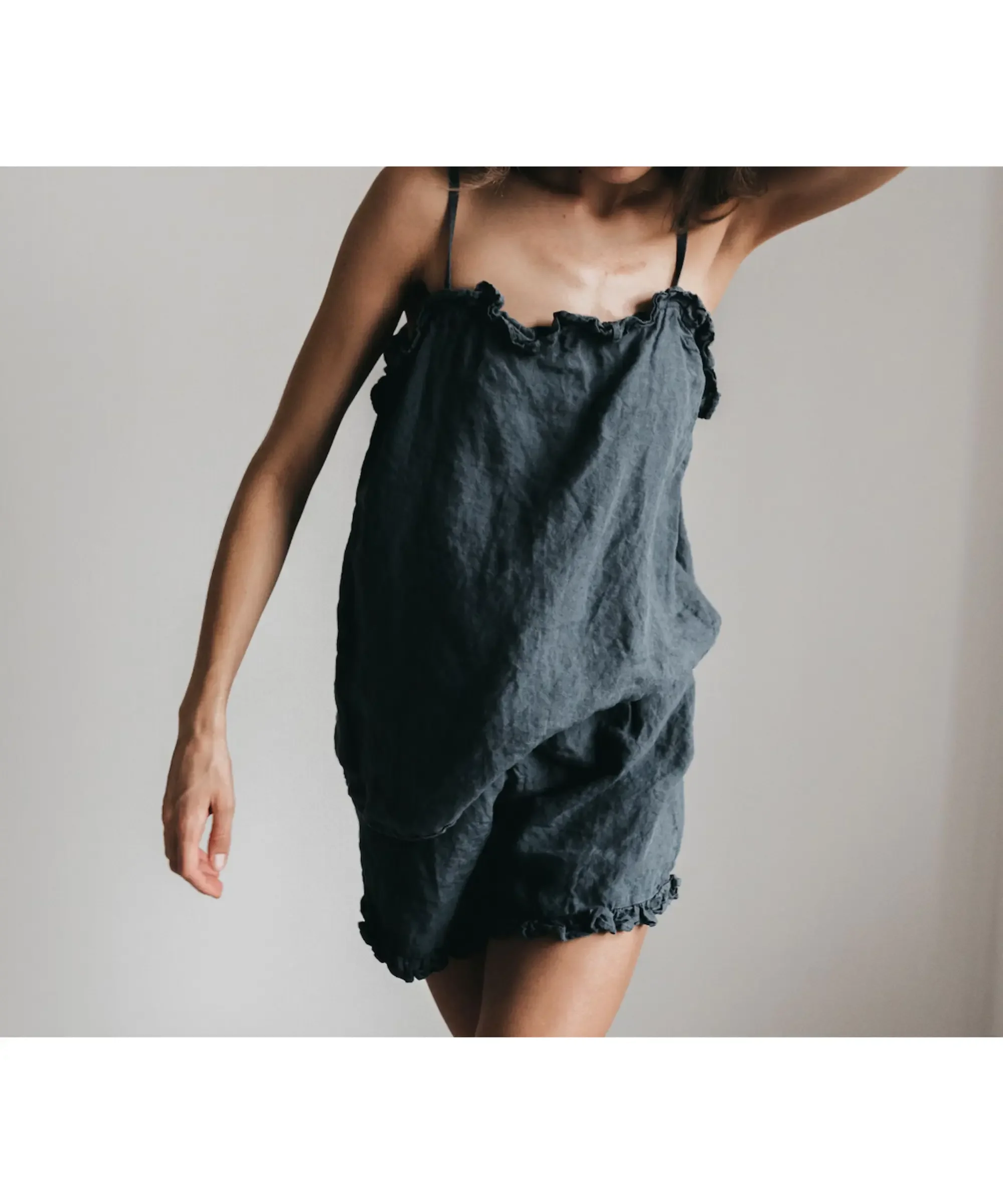 Dark gray linen pajama set featuring a relaxed-fit camisole top with delicate ruffle trim and matching loose shorts, worn by a woman in a minimalist, soft-lit setting.