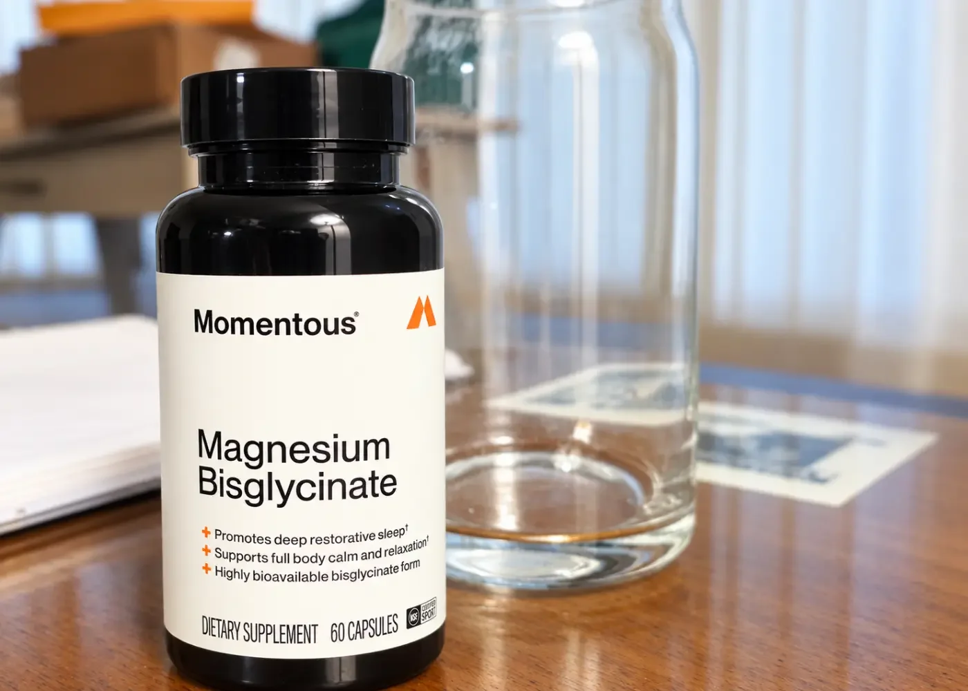 Momentous Magnesium Bisglycinate bottle with 60 capsules on a wooden table beside a clear glass water bottle.