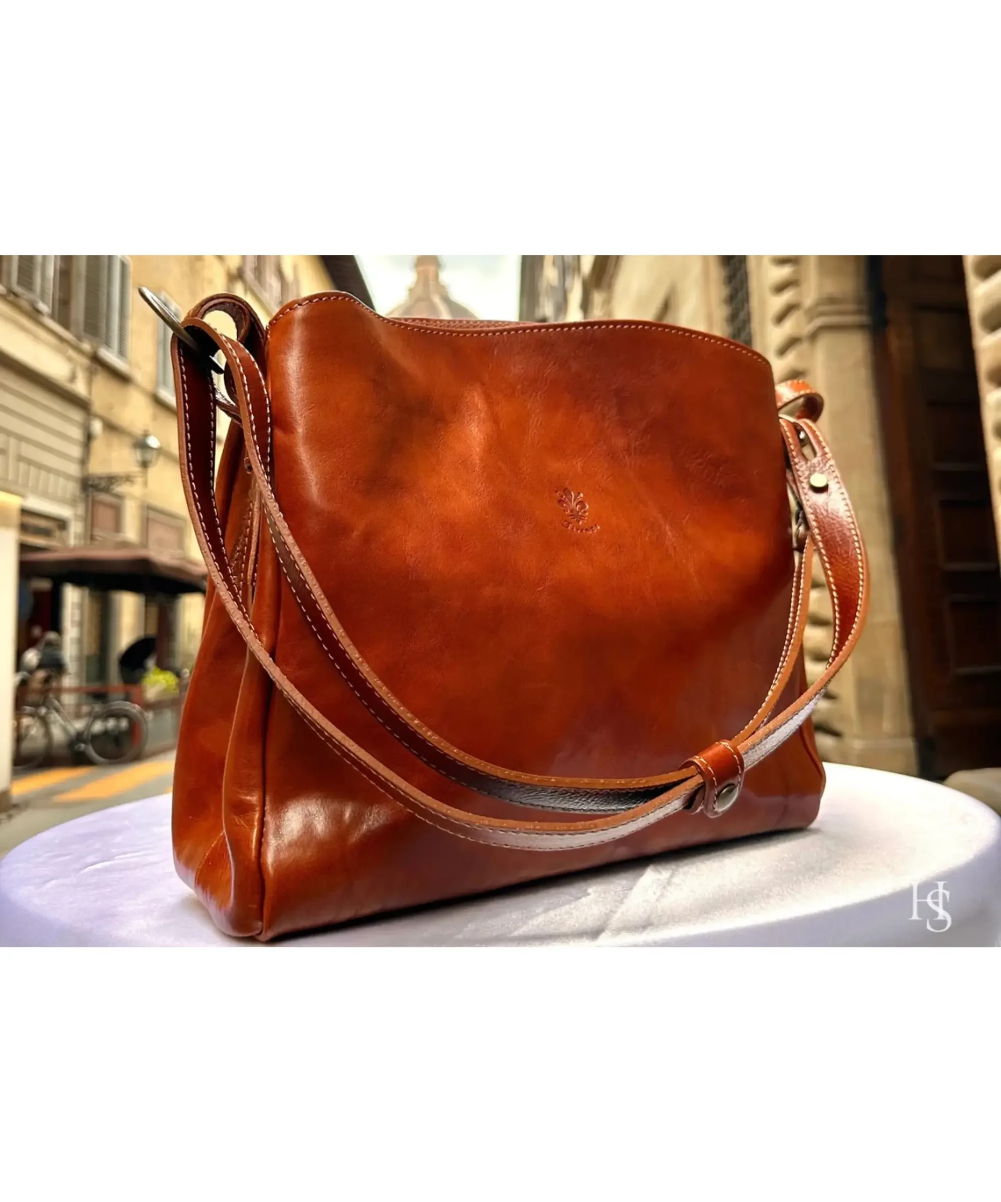 Italian handmade leather crossbody bag in rich brown leather with shoulder strap, elegant women’s bag displayed on a table in a Florence street setting.