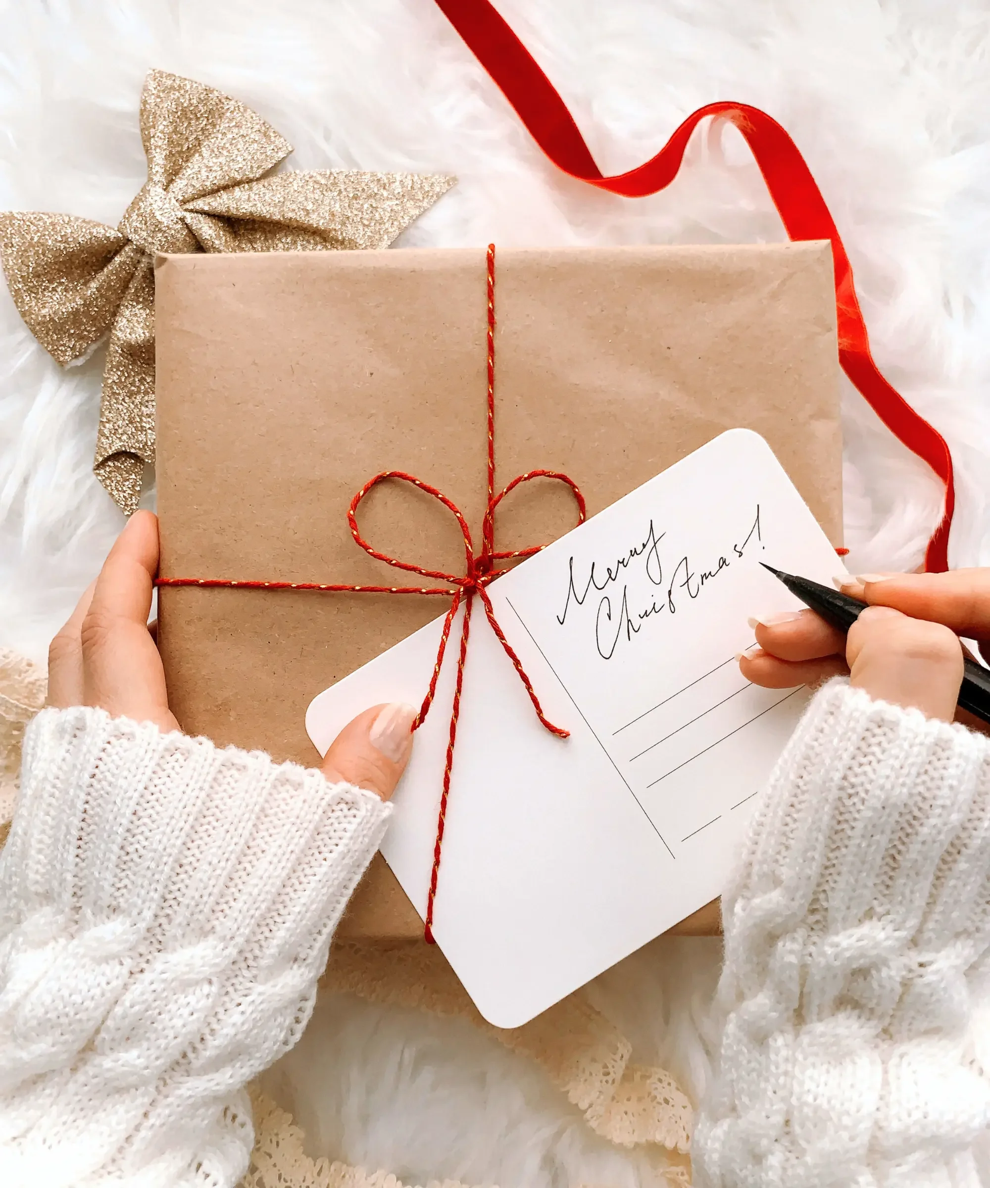 Hands in a white knit sweater wrapping a brown paper Christmas gift with red string and a handwritten “Merry Christmas” gift tag on a soft white background, minimalist holiday wellness gift aesthetic.
