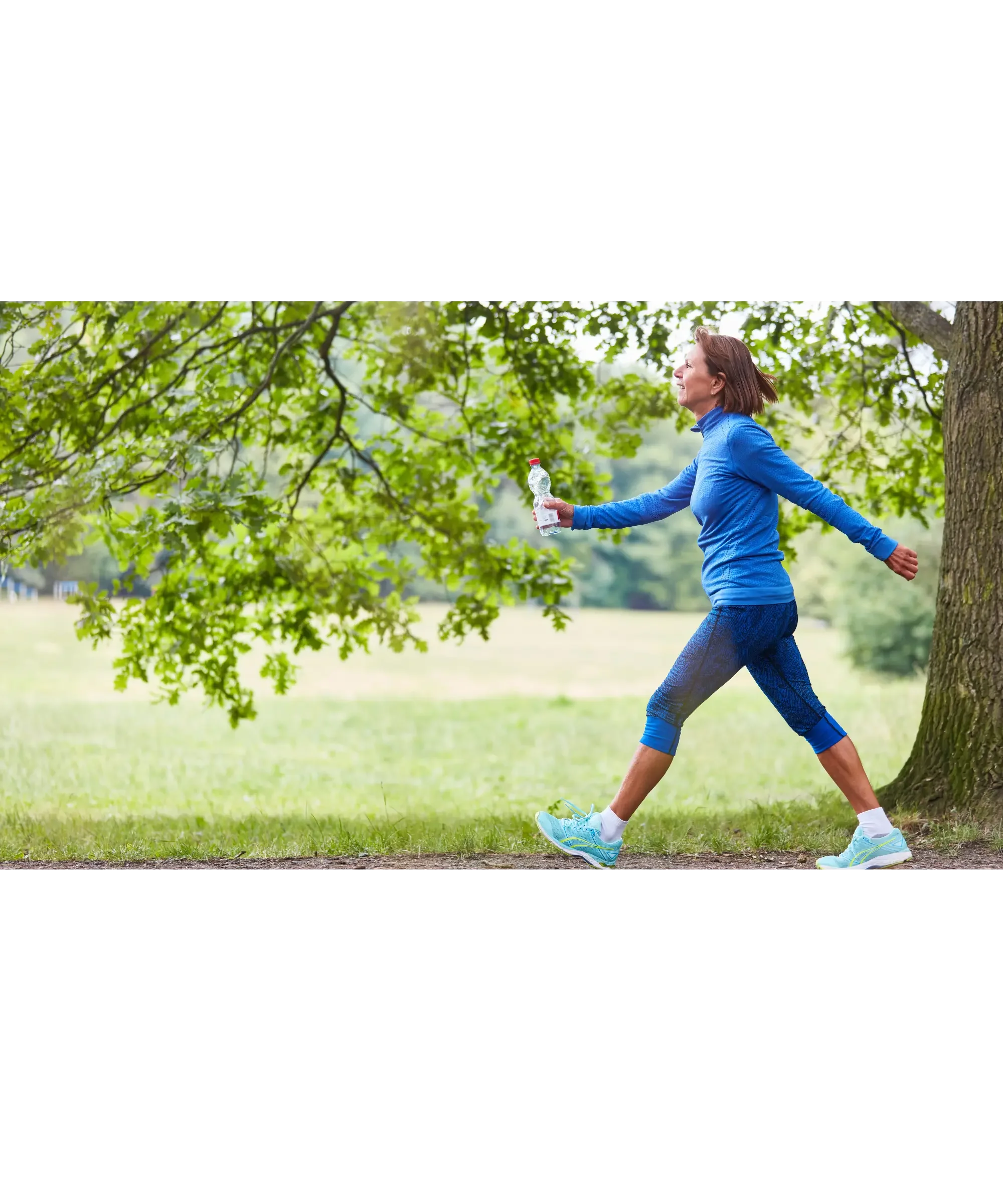 Woman in athletic wear walking briskly through a green park, holding a water bottle and enjoying an outdoor fitness routine.