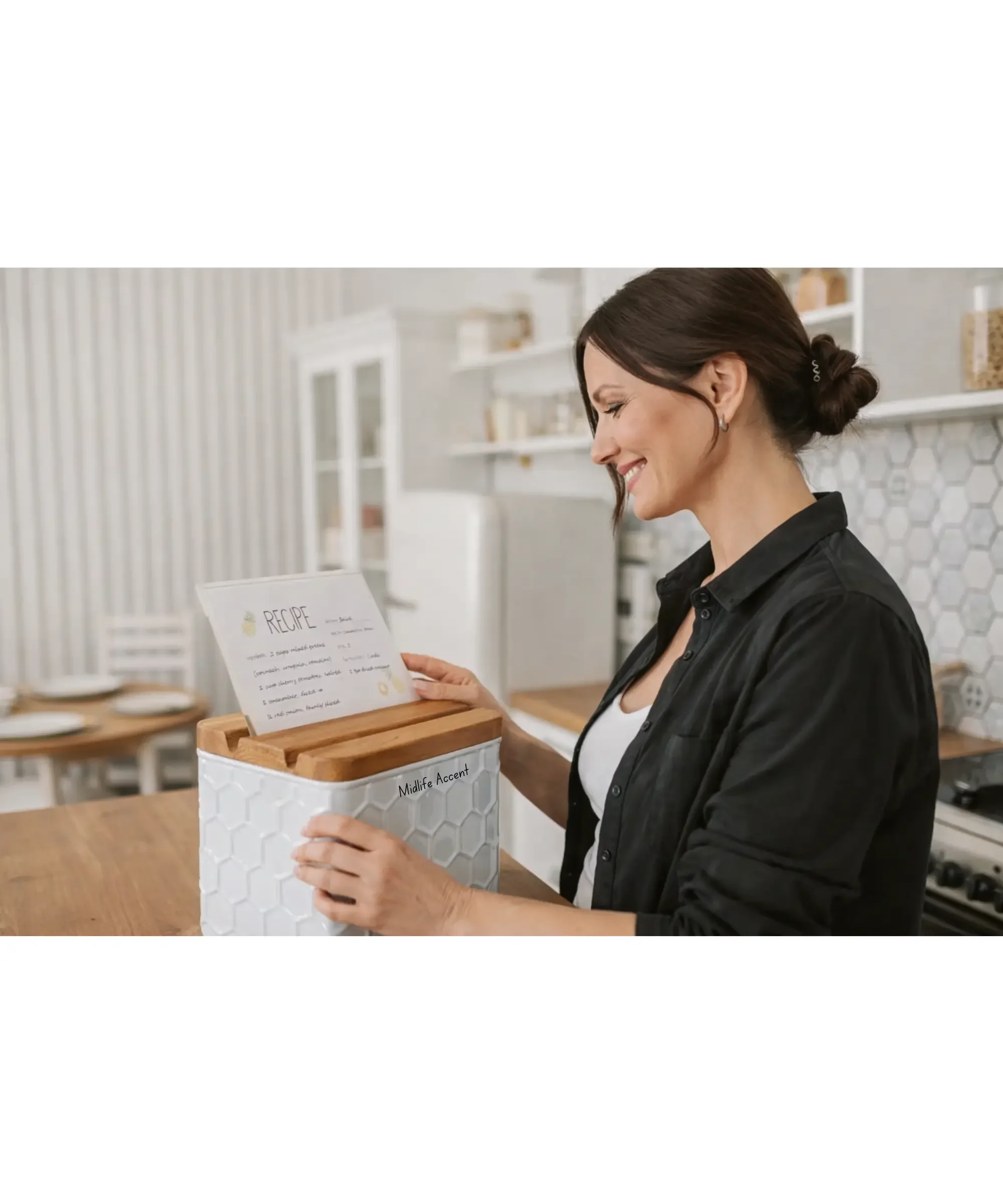 Smiling woman in her mid-40s standing in a bright kitchen, reading a recipe card from a white metal recipe box with a wooden lid on the counter.