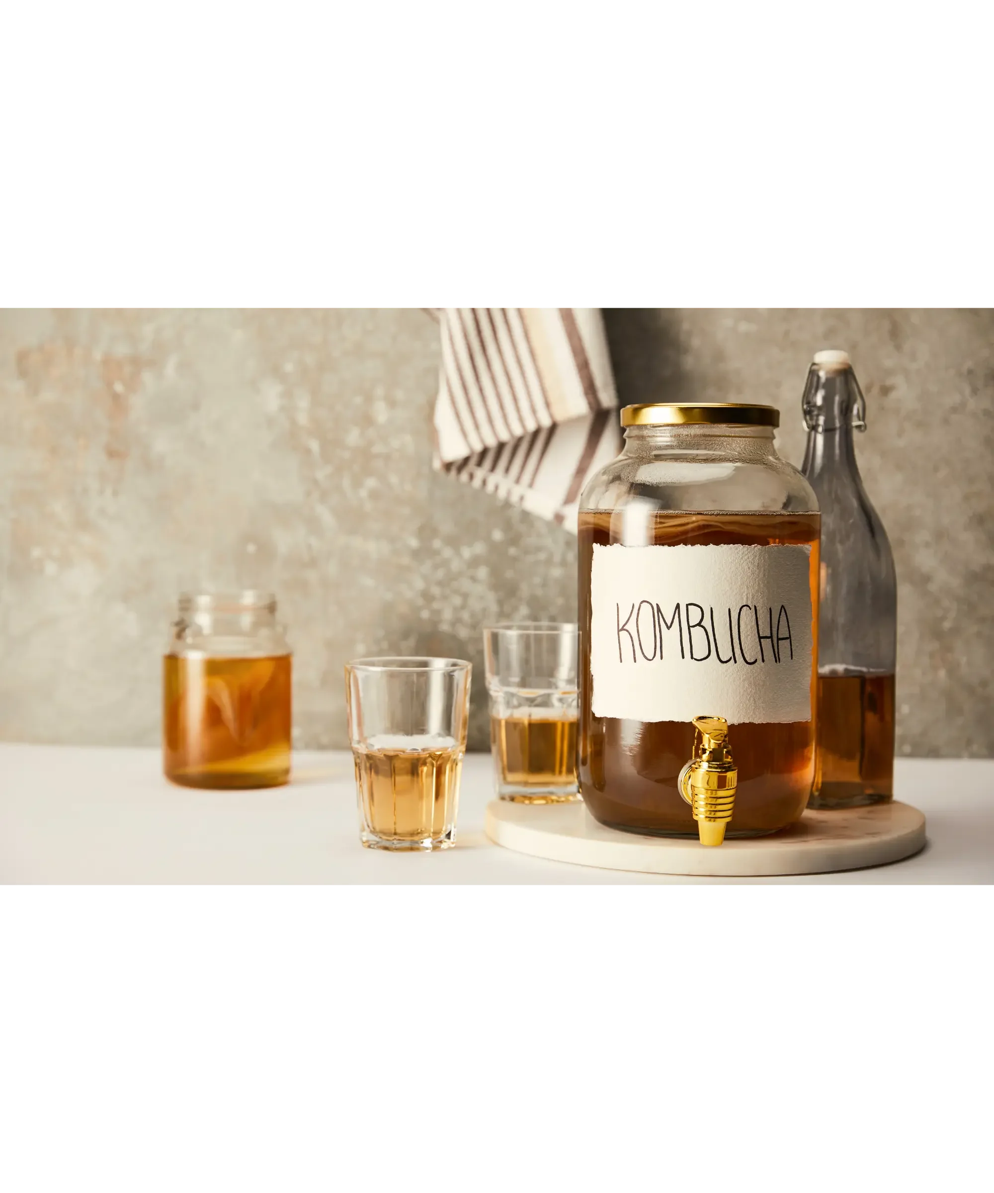 Glass jar labeled “kombucha” with a spigot, surrounded by bottles and glasses of fermented tea on a kitchen counter.
