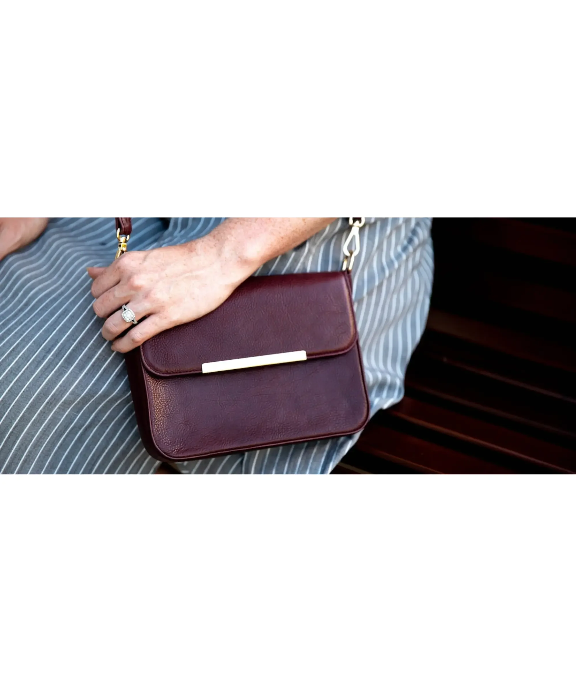 Small burgundy leather crossbody bag with gold accent bar, worn over a striped dress, elegant structured mini shoulder purse for women.