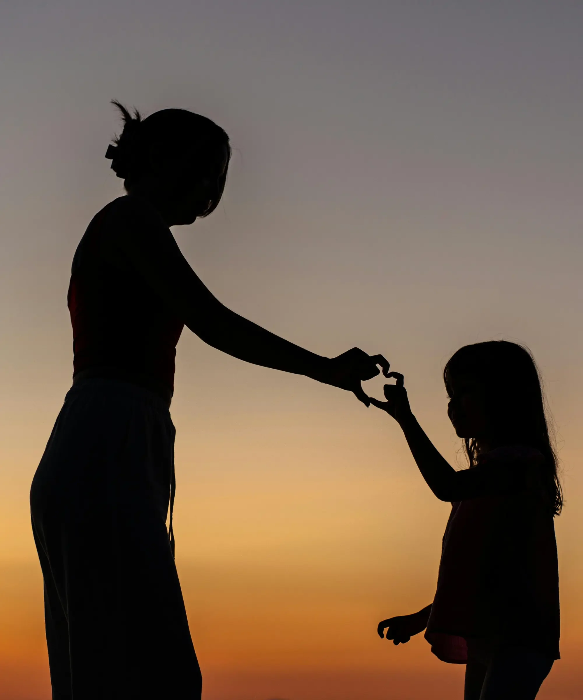 Silhouette of a mother and daughter at sunset forming a heart shape with their hands, symbolizing love, resilience, and the beauty of imperfect lives.
