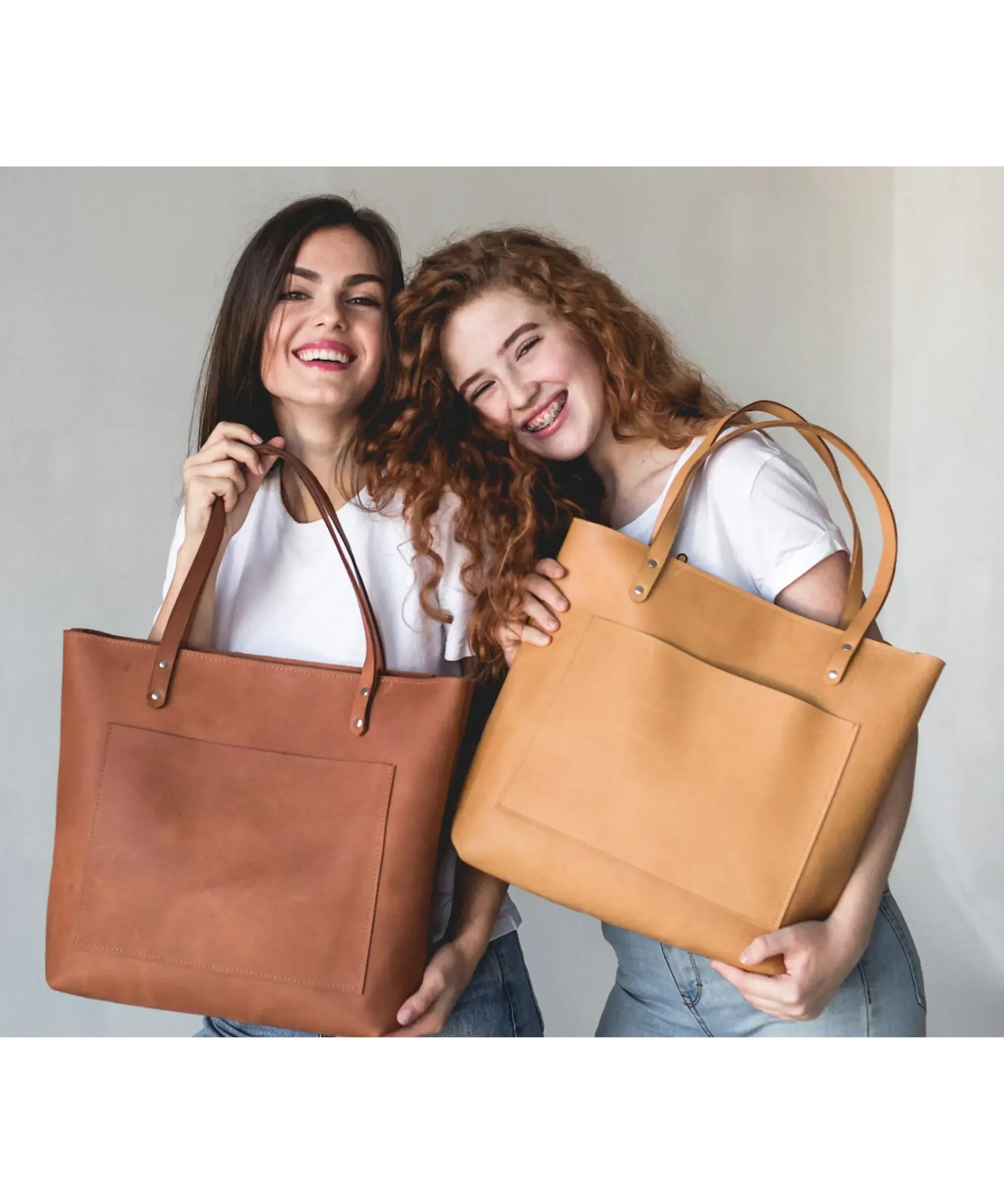 Two women smiling while holding large handmade leather tote bags in caramel and tan colors, stylish leather bags for women with shoulder straps.
