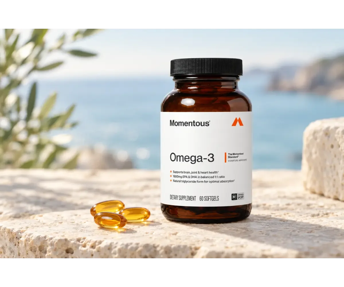 Momentous Omega-3 supplement bottle with three amber softgels displayed on pale stone near a softly blurred blue coastline.