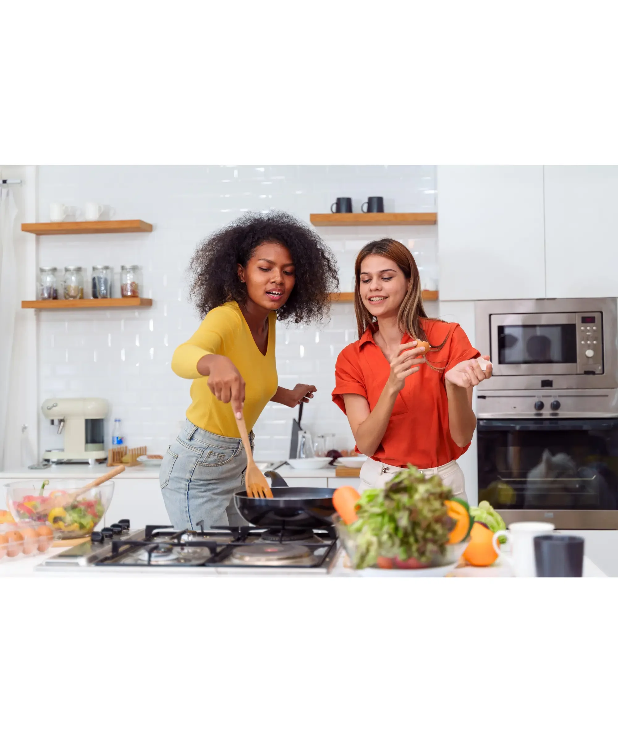 Two women cooking together in a bright modern kitchen, stirring food in a pan on the stove with fresh vegetables on the counter, representing everyday home cooking, healthy meals, and modern kitchen lifestyle.