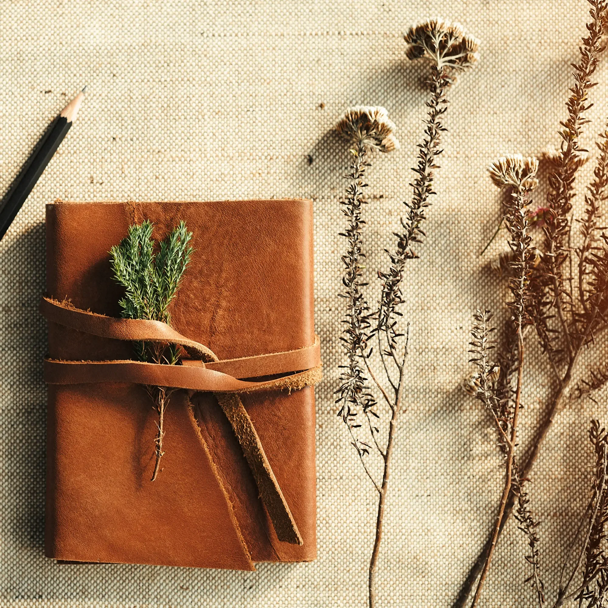 Minimalist flat lay of a brown leather-bound journal tied with a leather strap and a sprig of greenery, placed on natural linen fabric with dried wildflowers and a pencil, calm wellness and mindfulness aesthetic.