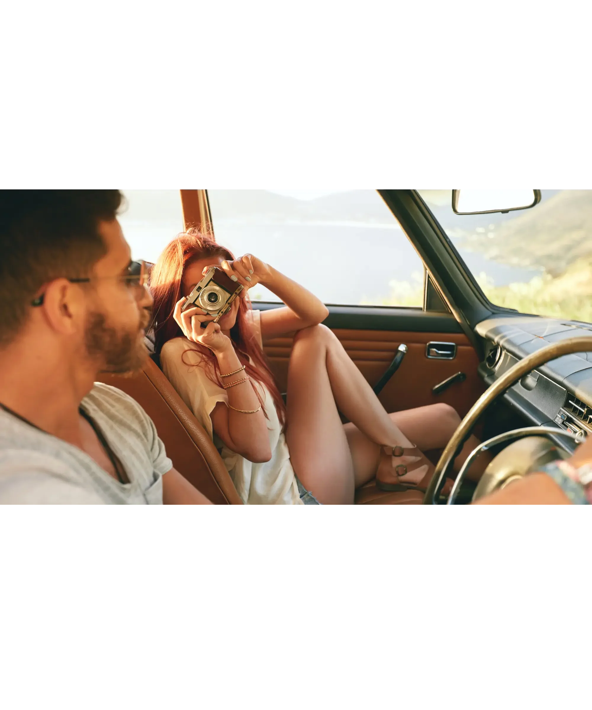 Woman in a car taking a photo with a vintage camera while sitting in the passenger seat, with a man driving beside her during a scenic road trip.