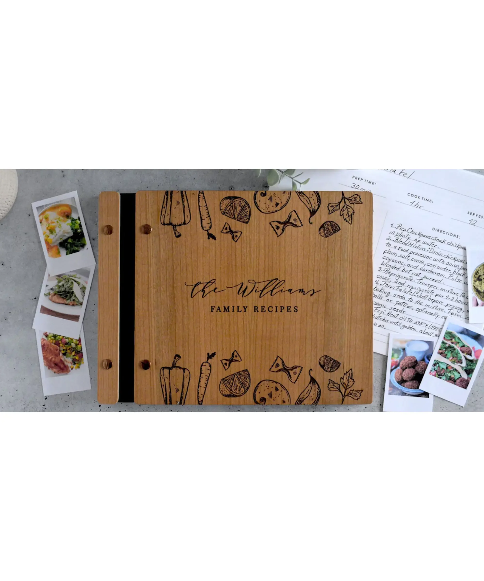 Personalized wooden family recipe book binder engraved with “The Williams Family Recipes,” surrounded by printed recipes and food photos on a kitchen table.