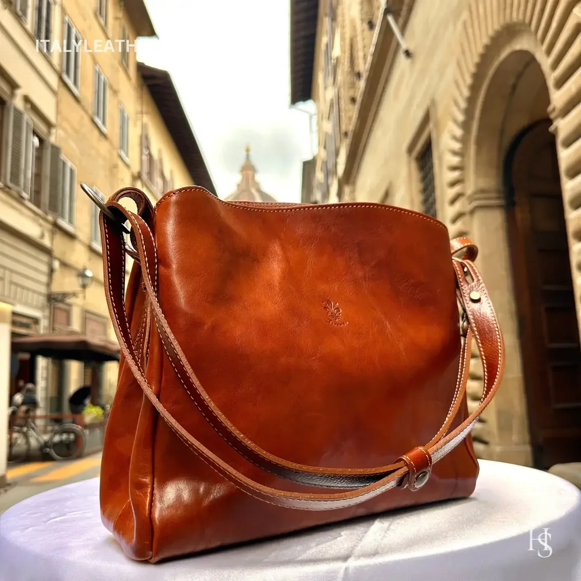 Italian handmade brown leather crossbody bag with adjustable strap, soft structured design, and classic everyday style, photographed on a European street backdrop