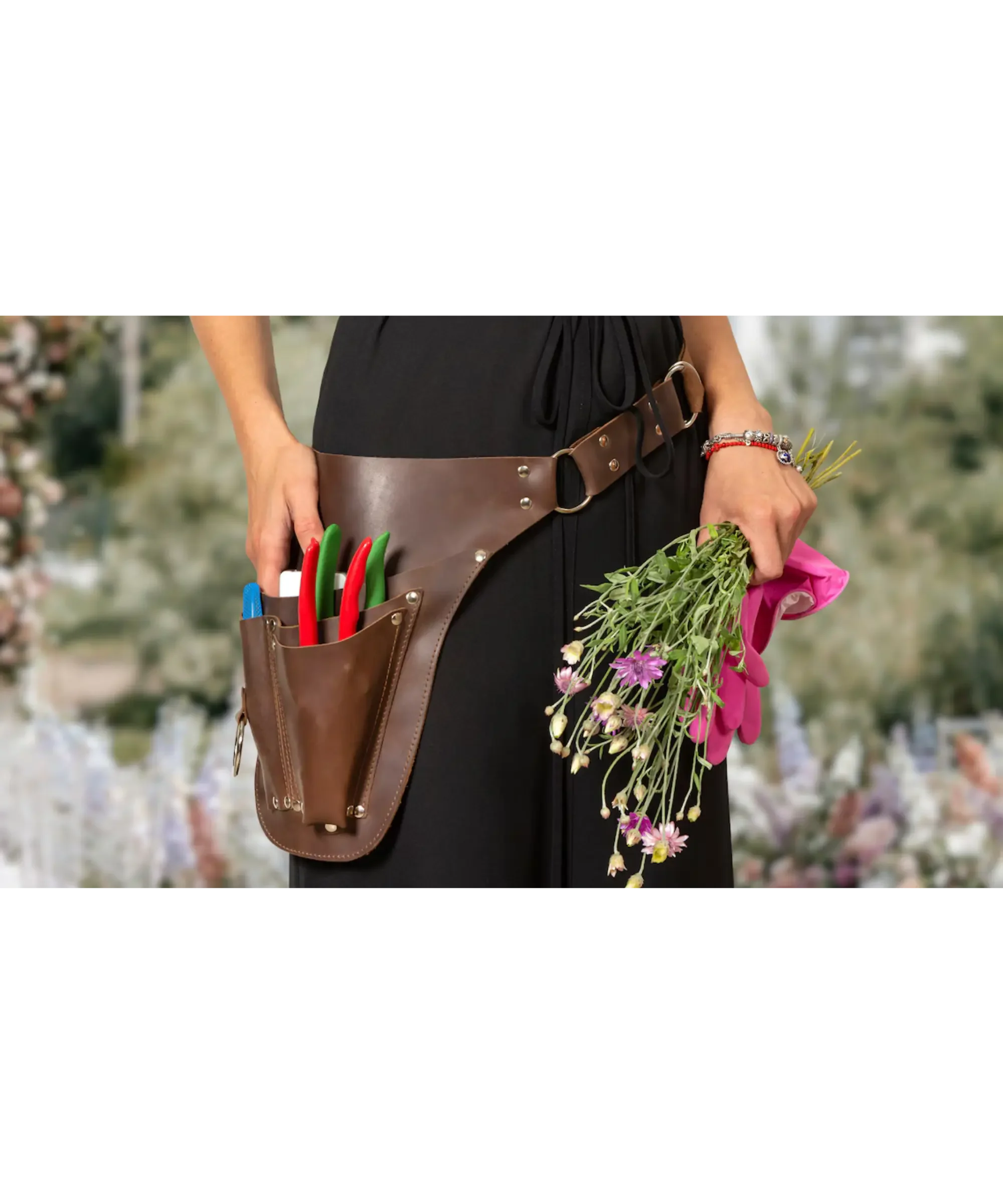 Person wearing a leather gardening apron with tools, holding flowers and gloves in a garden setting.