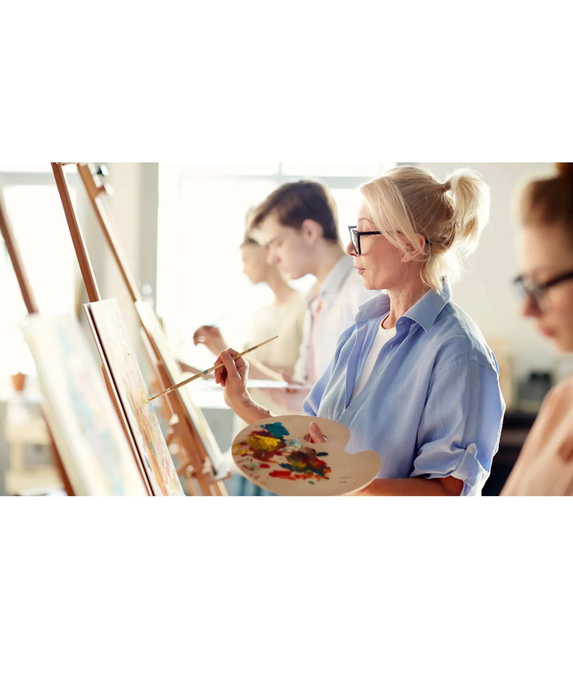 Older woman painting on a canvas during an art class, holding a palette and brush