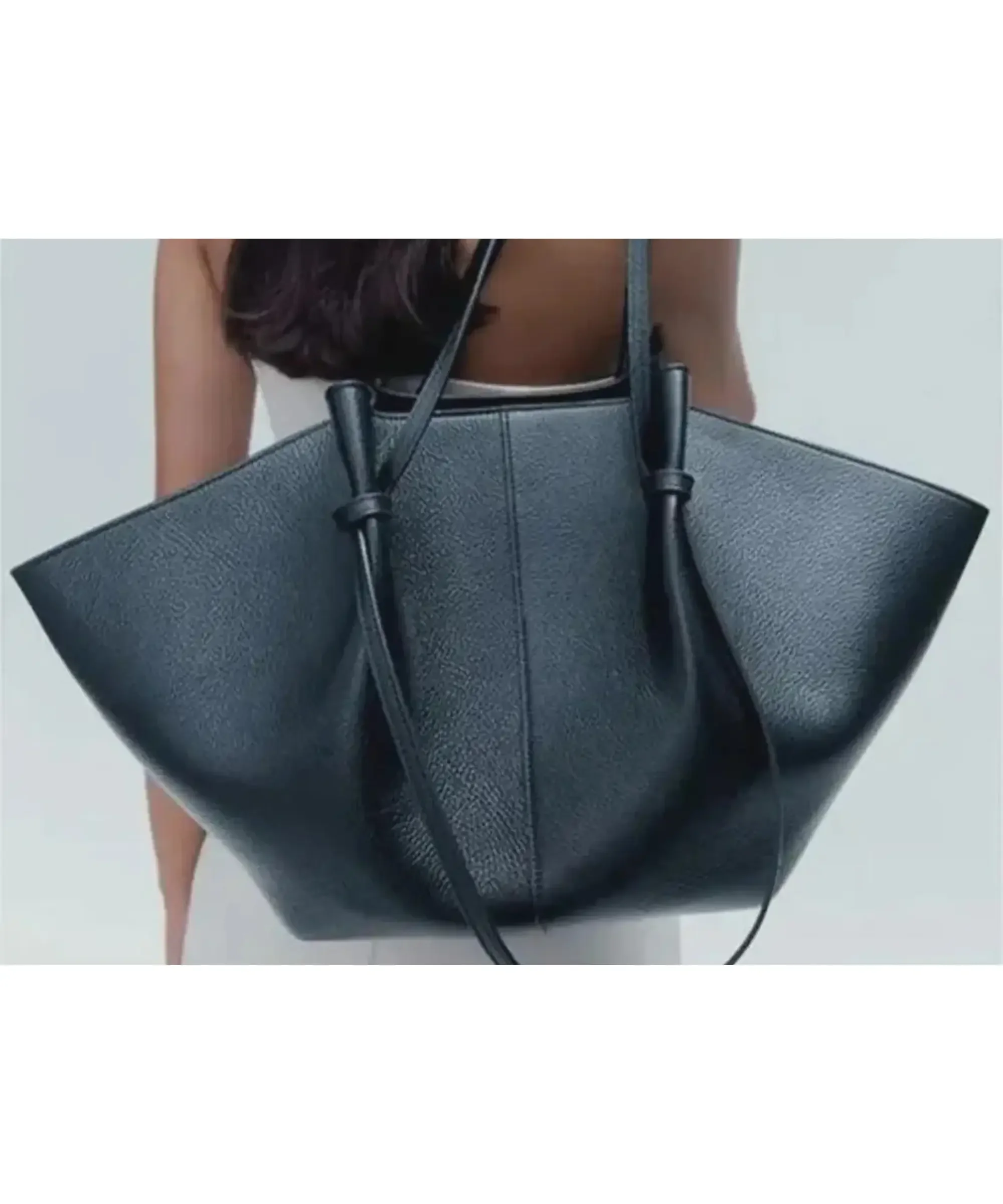 Large black handcrafted leather shoulder tote bag with wide structured sides, worn by a woman, elegant everyday leather handbag for commuting or work.